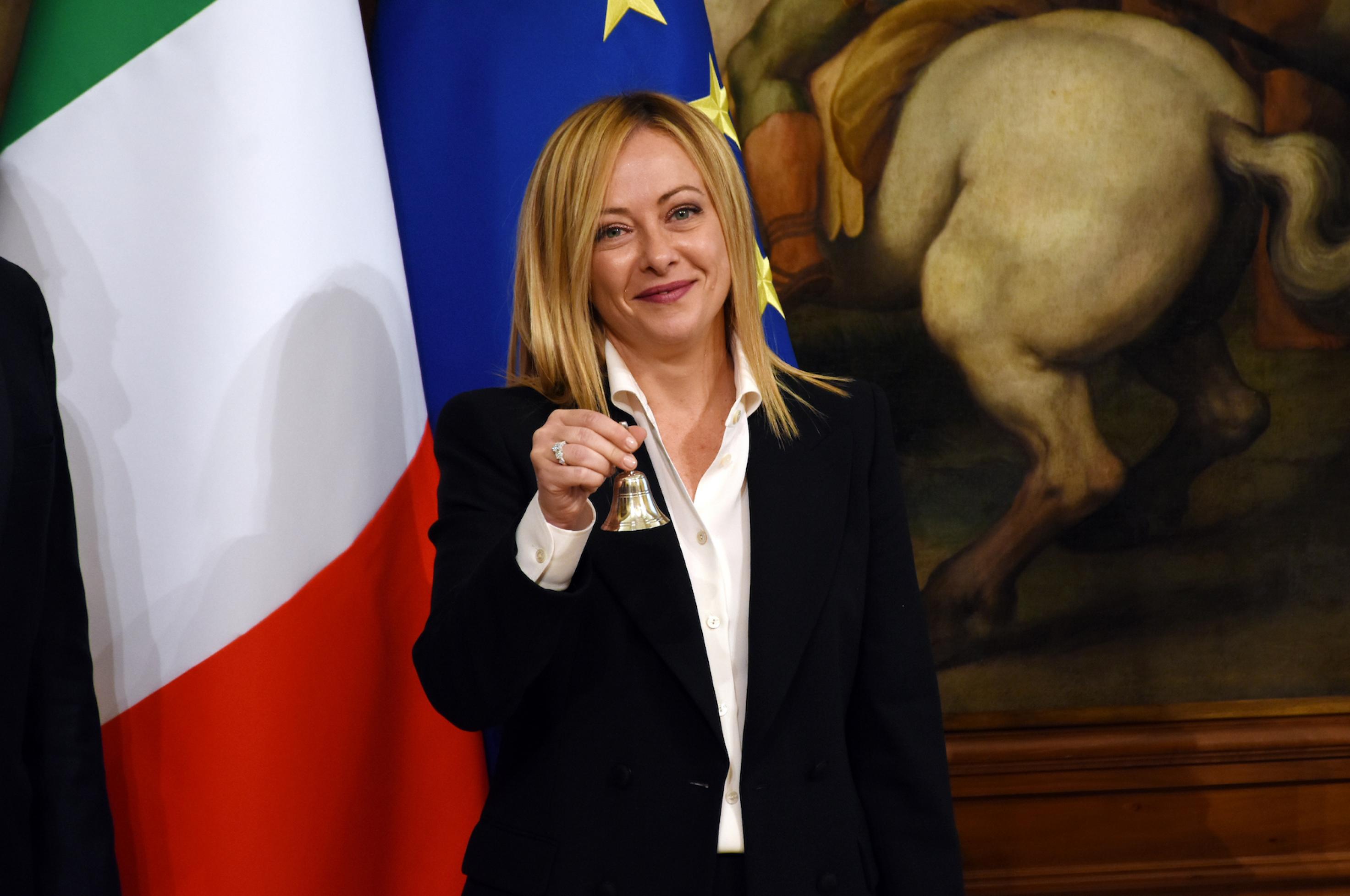 Die italienische Ministerpräsidentin im Halbportrait, sie trägt eine dunkle Anzug-oder Kostümjacke zu einer weißen Bluse. Links von ihr sind Ausschnitte der italienischen und der europäischen Flagge zu sehen, im Hintergrund ein kleiner Ausschnitt aus einem Ölgemälde: das gut bemuskelte Hinterteil eines Pferdes, das sich mit den Vorderbeinen etwas aufgerichtet hat.