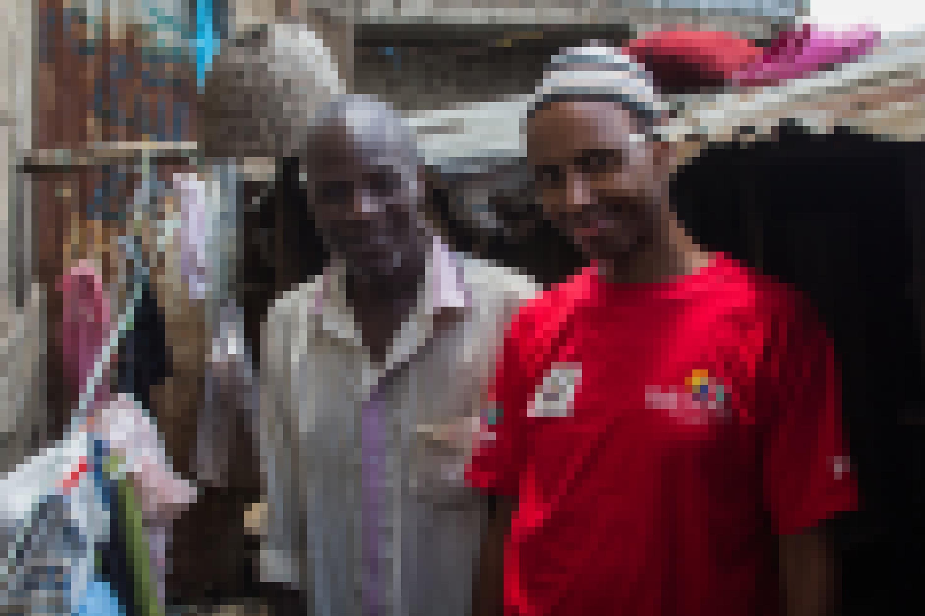 Jacob Omondi und Salat Hamud stehen nebeneinander am Eingang einer sehr schmalen Gasse, rechts hinter ihnen stehen niedrige Wellblechhütten, links eine hohe Mauer.