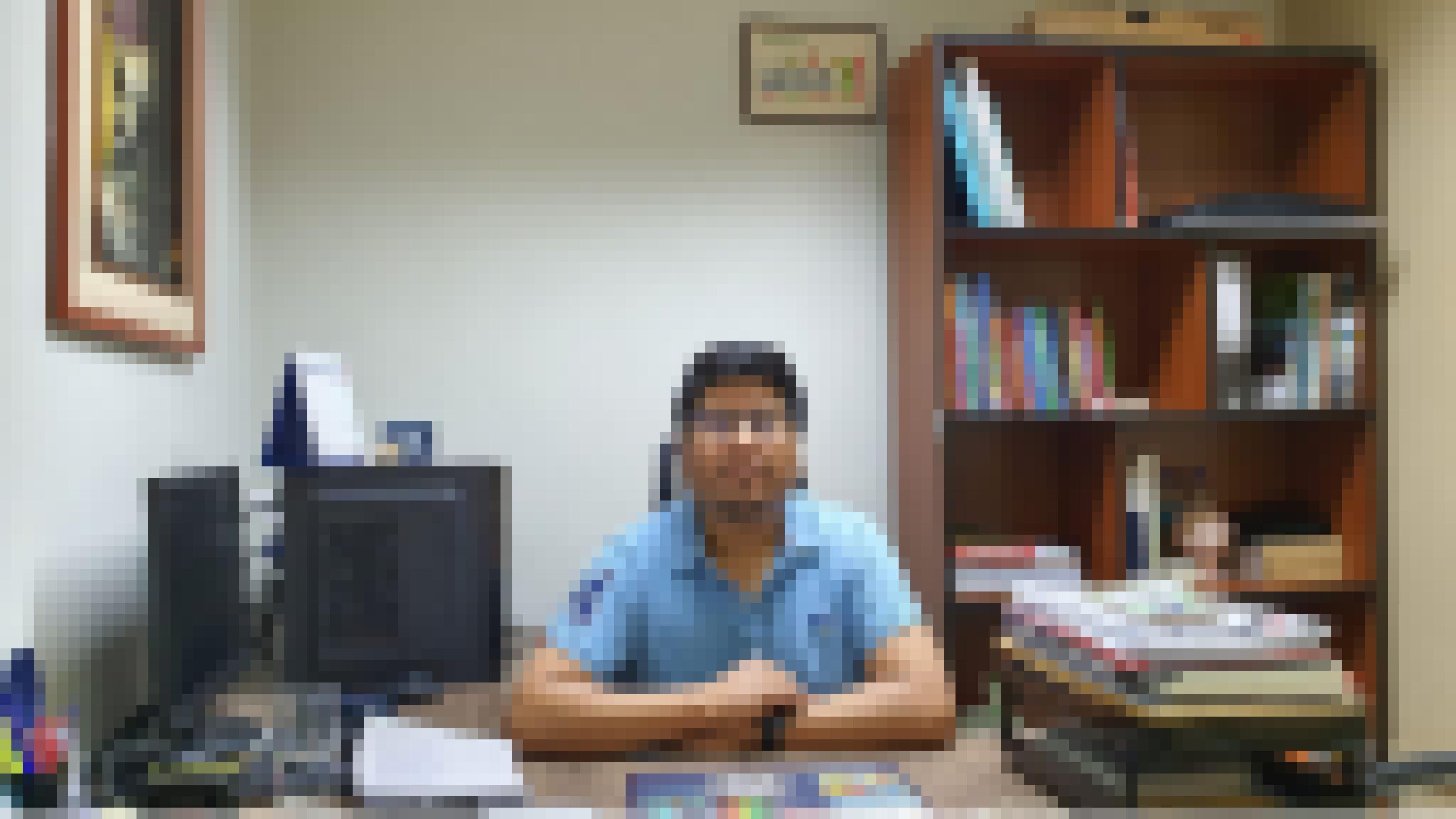 Mann Mitte 30 sitzt hinter einem Schreibtisch. Er trägt ein hellblaues Polohemd mit kurzen Ärmeln. Auf dem Schreibtisch Computer, Papierstapel, Wasserglas, Schreibzeug, links ein Druck an der Wand und hinter ihm ein Buchregal, in dem noch Platz ist.