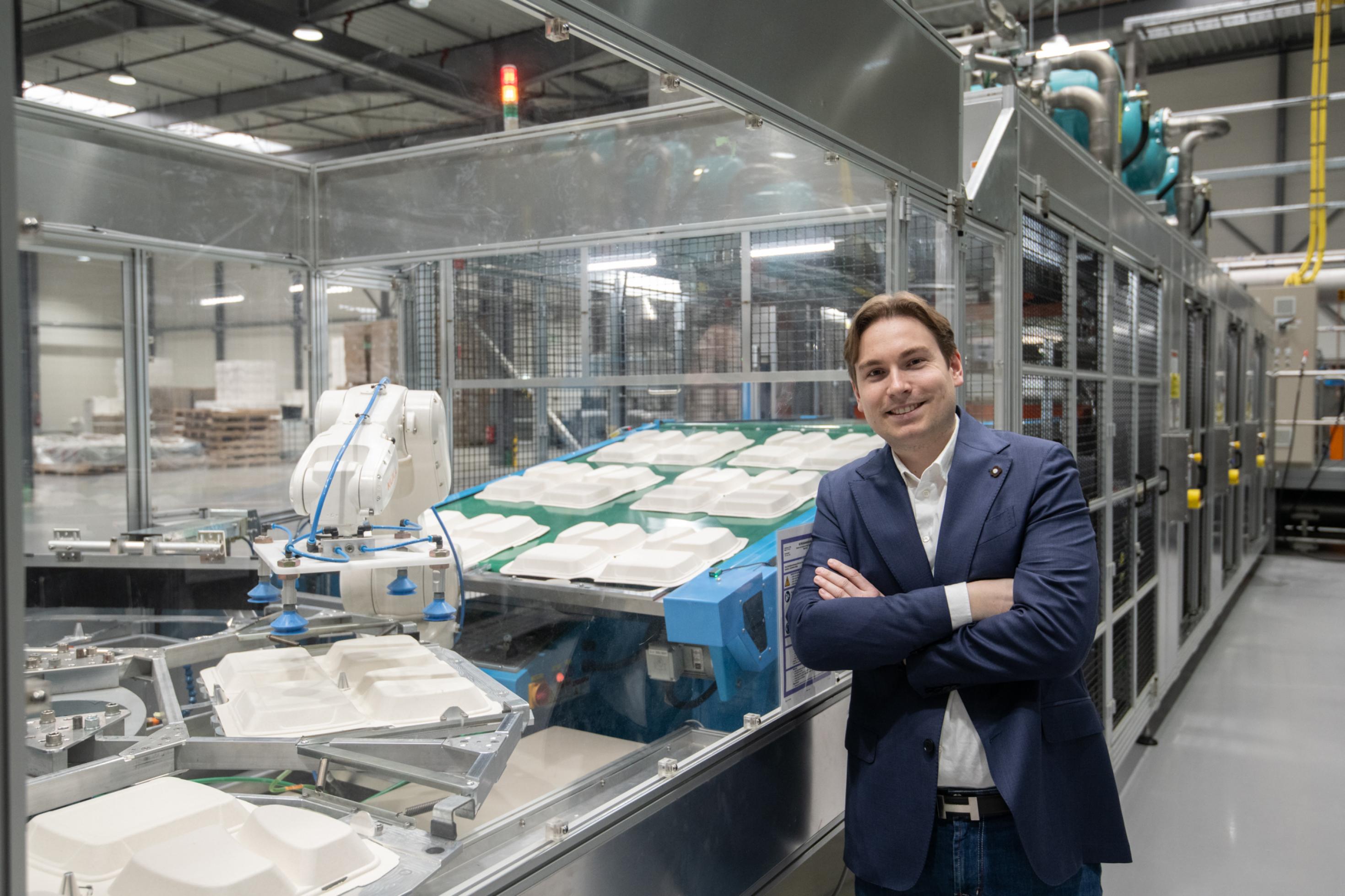 Ein junger Mann in Jeans und Jackett steht vor einer Anlage, die Verpackungen für Fastfood produziert.