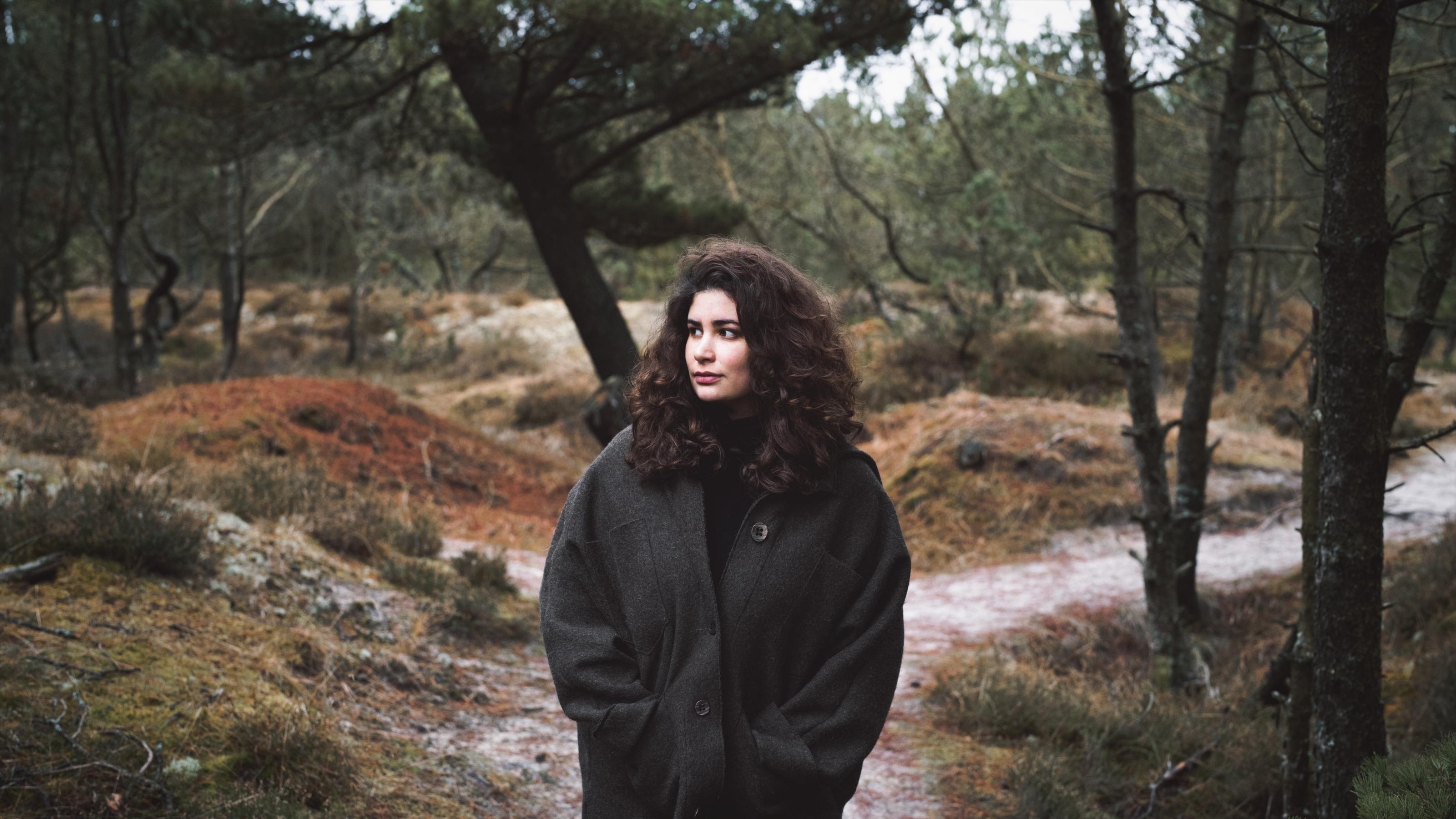Zu sehen ist eine Frau mit dunklen, langen Locken – die Autorin Jasmin Schreiber -, die auf einem Waldweg steht. Um sie herum stehen Bäume.
