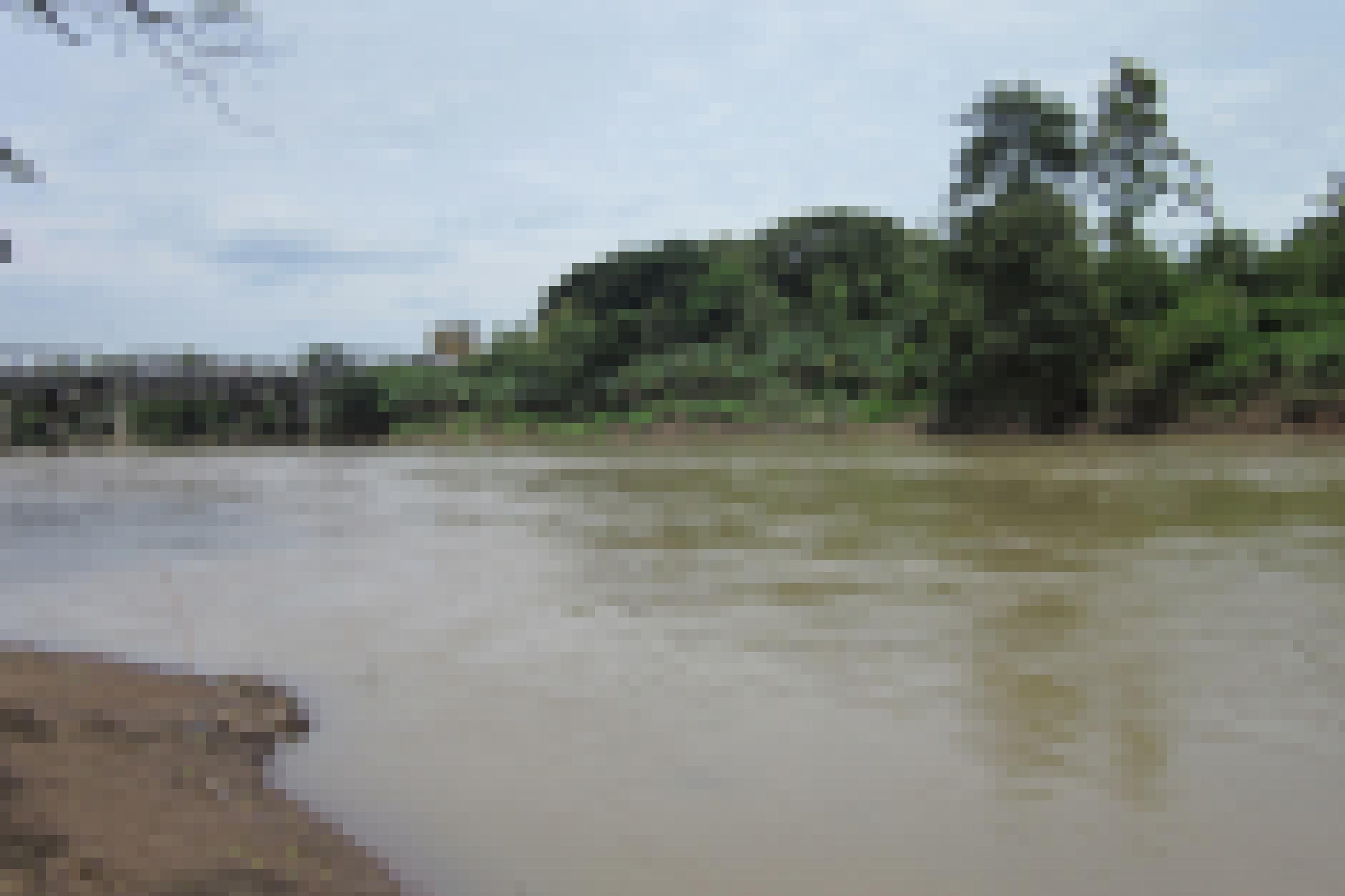 Das Bild zeigt den Blick auf einen Fluss, dessen Wasser eine trübe, grünbraune Farbe aufweist. An den Ufern schmale braune Sandstreifen, auf der rechten Seite schließen sich dem Streifen grüne Büsche und Bäume an. Links im Hintergrund eine Stahlbrücke, die über den Fluss führt.
