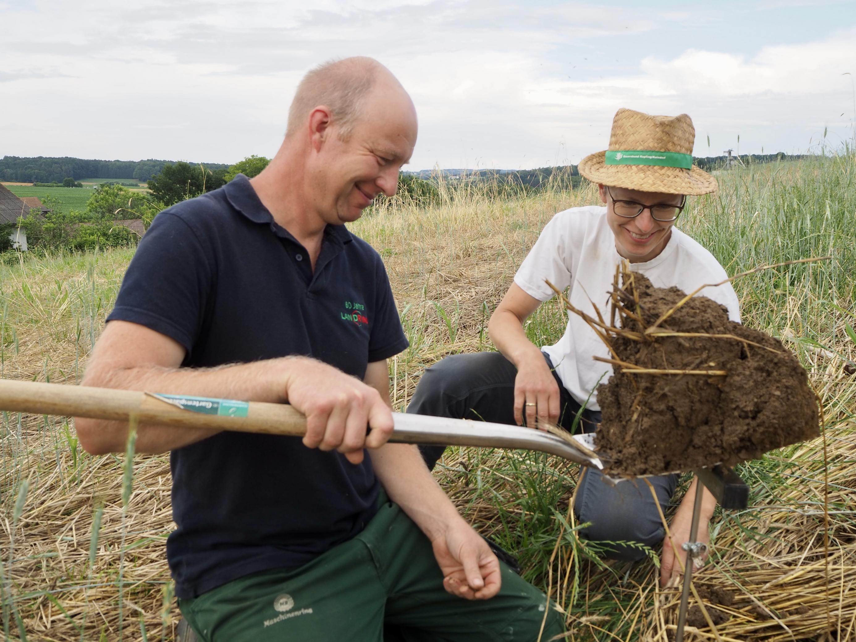 Die Landwirte Martina und Johann Höfler. Er betrachtet zufrieden einen Spatenaushub mit Ackererde. Sie trägt einen Strohhut und lächelt