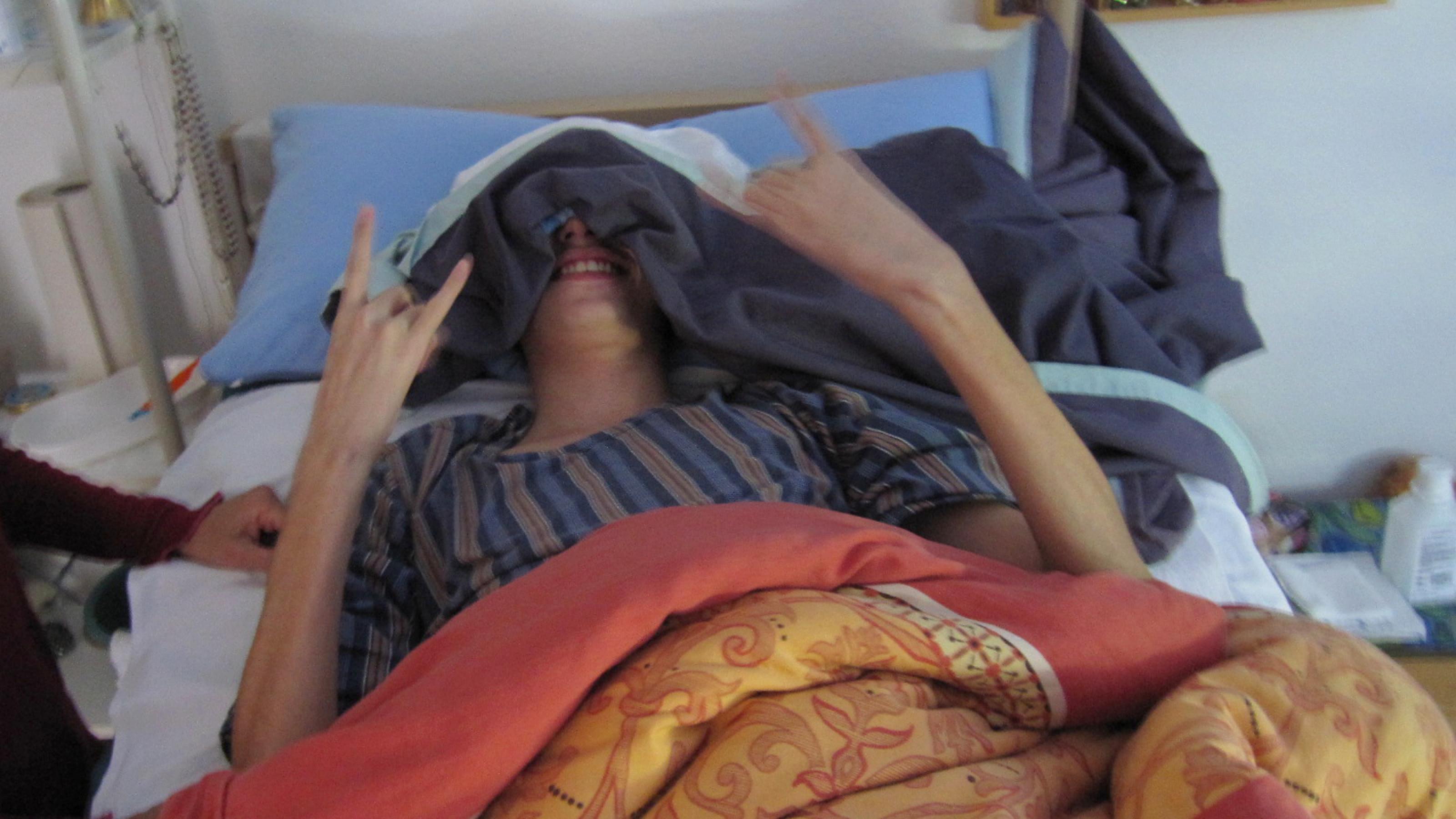 Der schwer an ME/CFS erkrankte Jonas liegt in seinem Pflegebett, das Gesicht unter einem Laken verdeckt, um die Augen vor Lichtreizen zu schützen. Nur der Mund ist frei und zeigt ein Lächeln, Jonas' Hände formen das Zeichen von Heavy-Metal-Fans mit ausgestrecktem Zeige- und kleinem Finger.