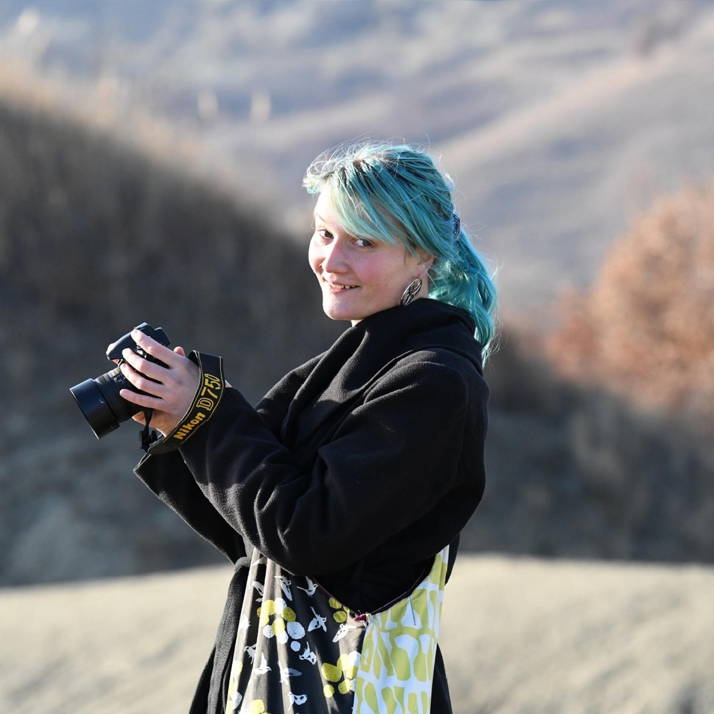 blauhaarige Frau mit Kamera in der Hand vor Bergen