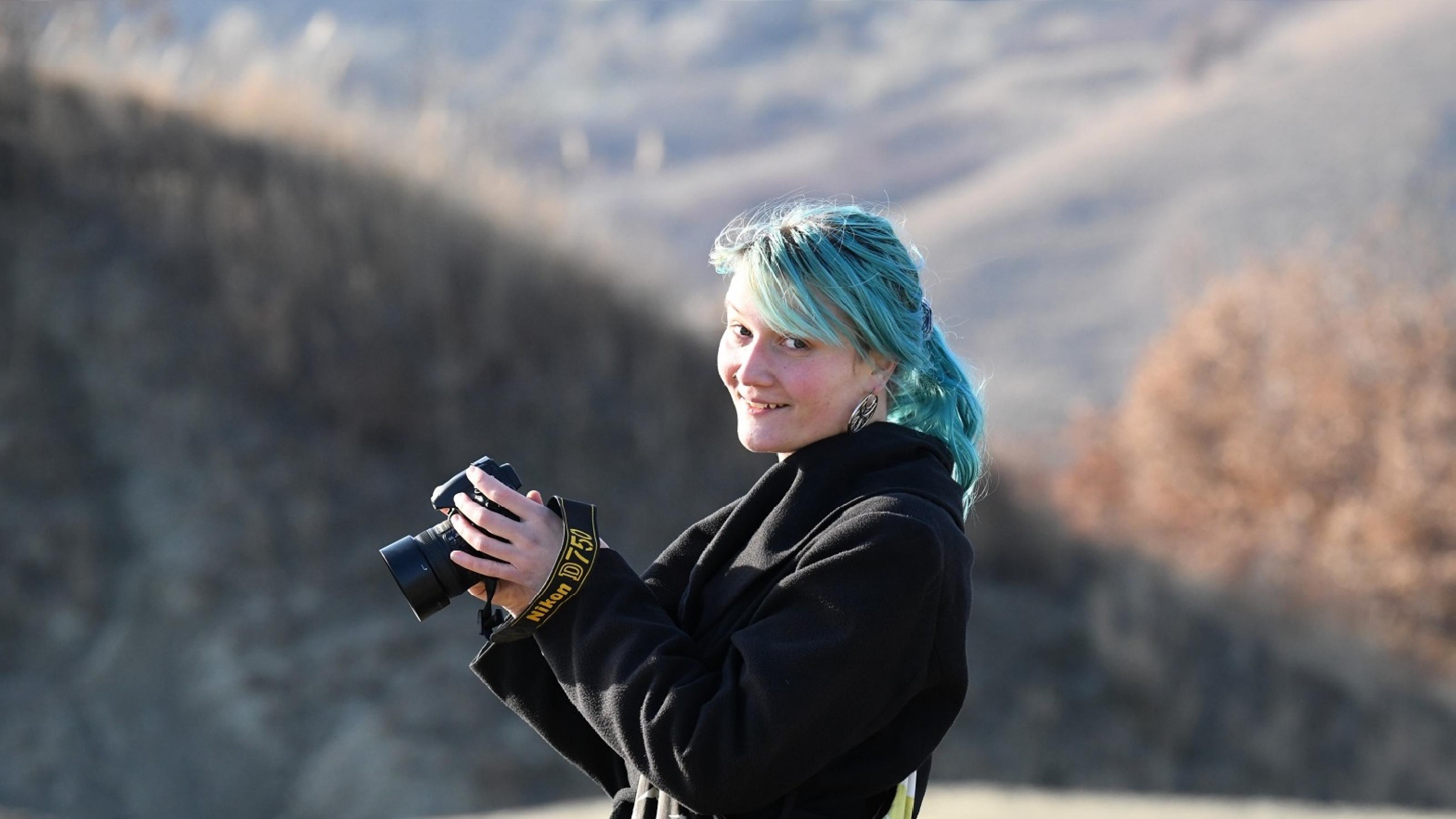 blauhaarige Frau mit Kamera in der Hand vor Bergen