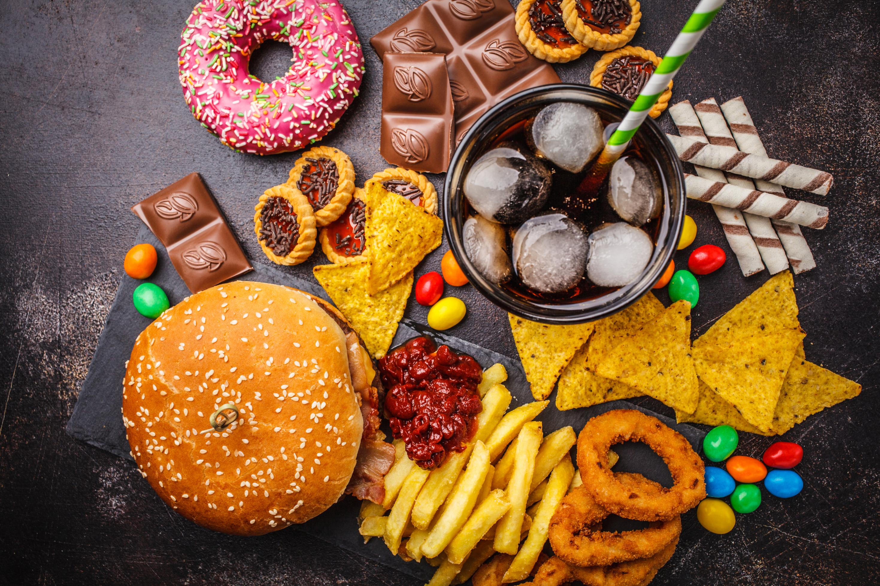 Draufsicht: Verschiedene Junk Food Produkte wie Burger, Süßigkeiten, Chips, Schokolade, Donuts, Soft Drinks – fotografiert auf schwarzer Tischplatte.