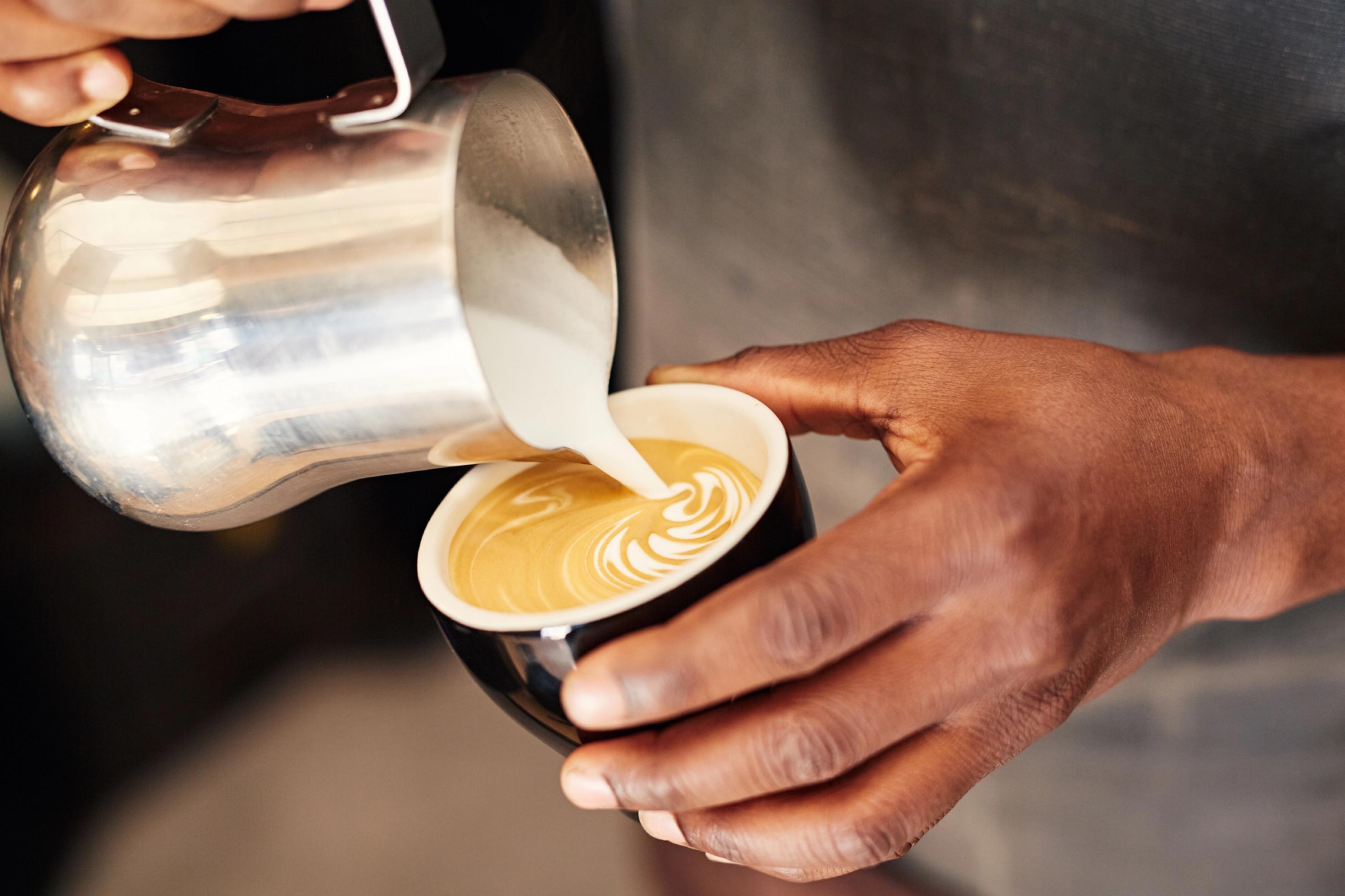 Nahaufnahme: Barista-Hand gießt Milchschaum in eine Tasse Kaffee.
