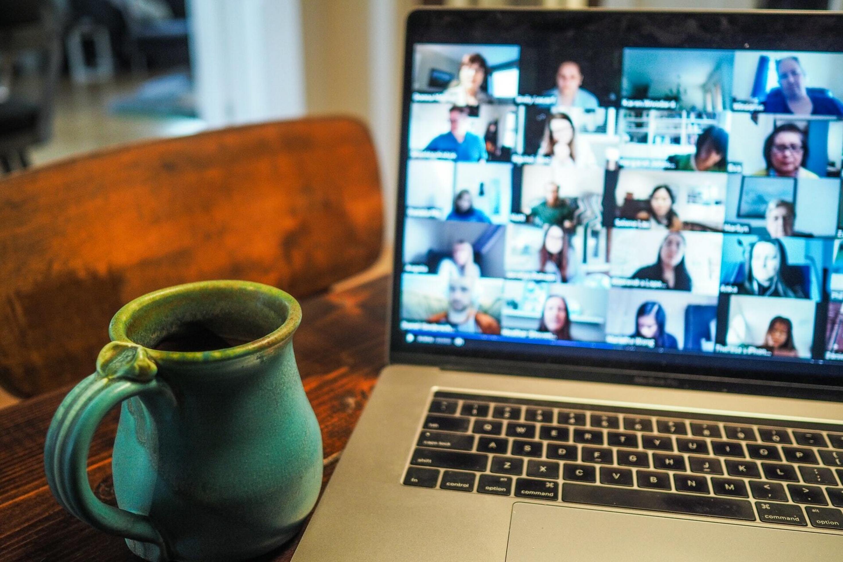 Eine Kaffeetasse steht auf einem Tisch vor einem Laptop. Auf dem Laptop findet eine Videokonferenz statt.