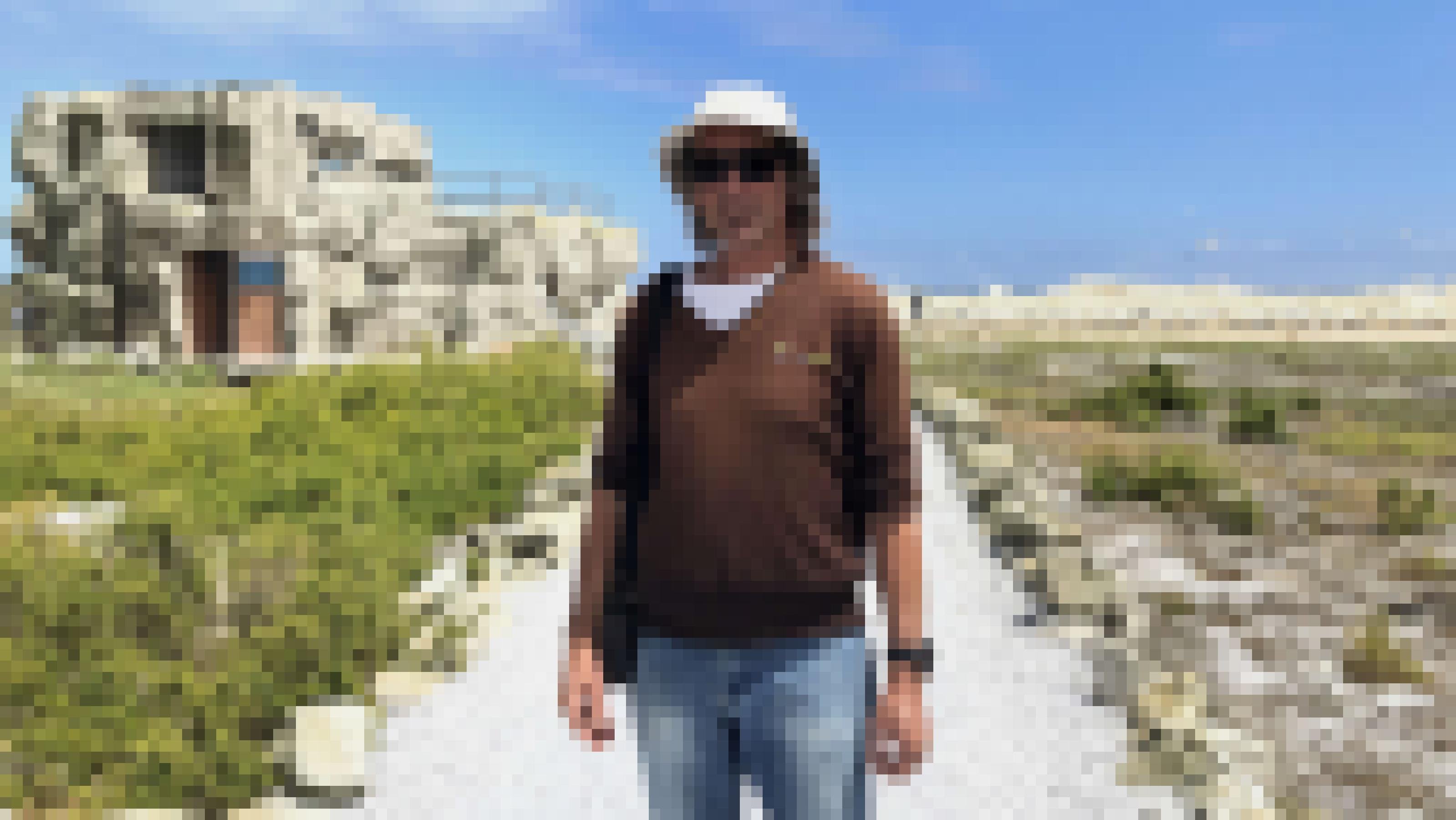 Ein Mann im braunen Pulli mit Mütze und Sonnenbrille steht auf einem Weg, der zu einem zweistöckigen Gebäude führt, das als Felsen getarnt ist.