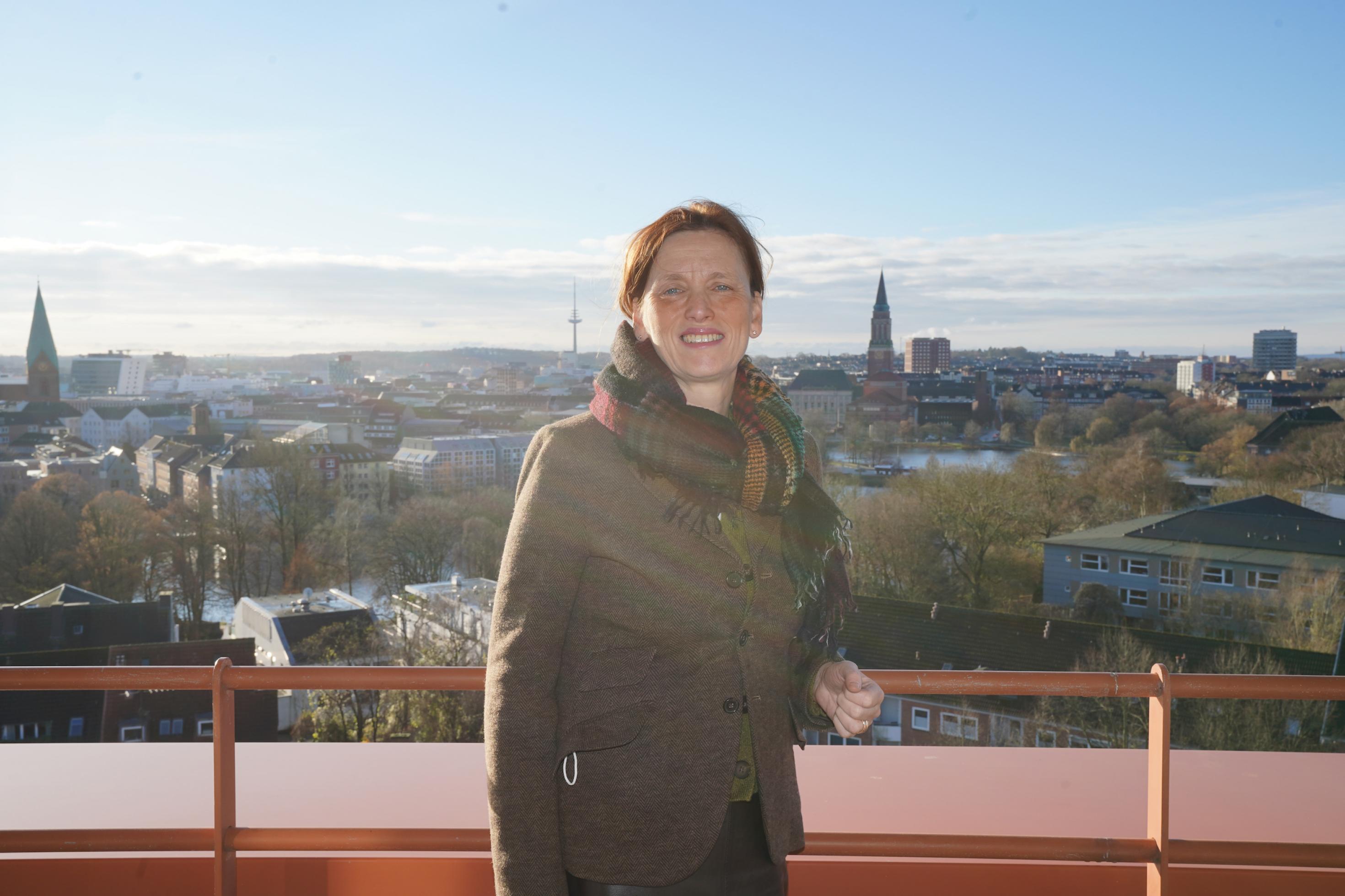 Karin Prien, eine Frau mit rotbraunen Haaren steht auf einer Dachterasse mit weitem Ausblick über Kiel. Man sieht zwei Kirchtürme.