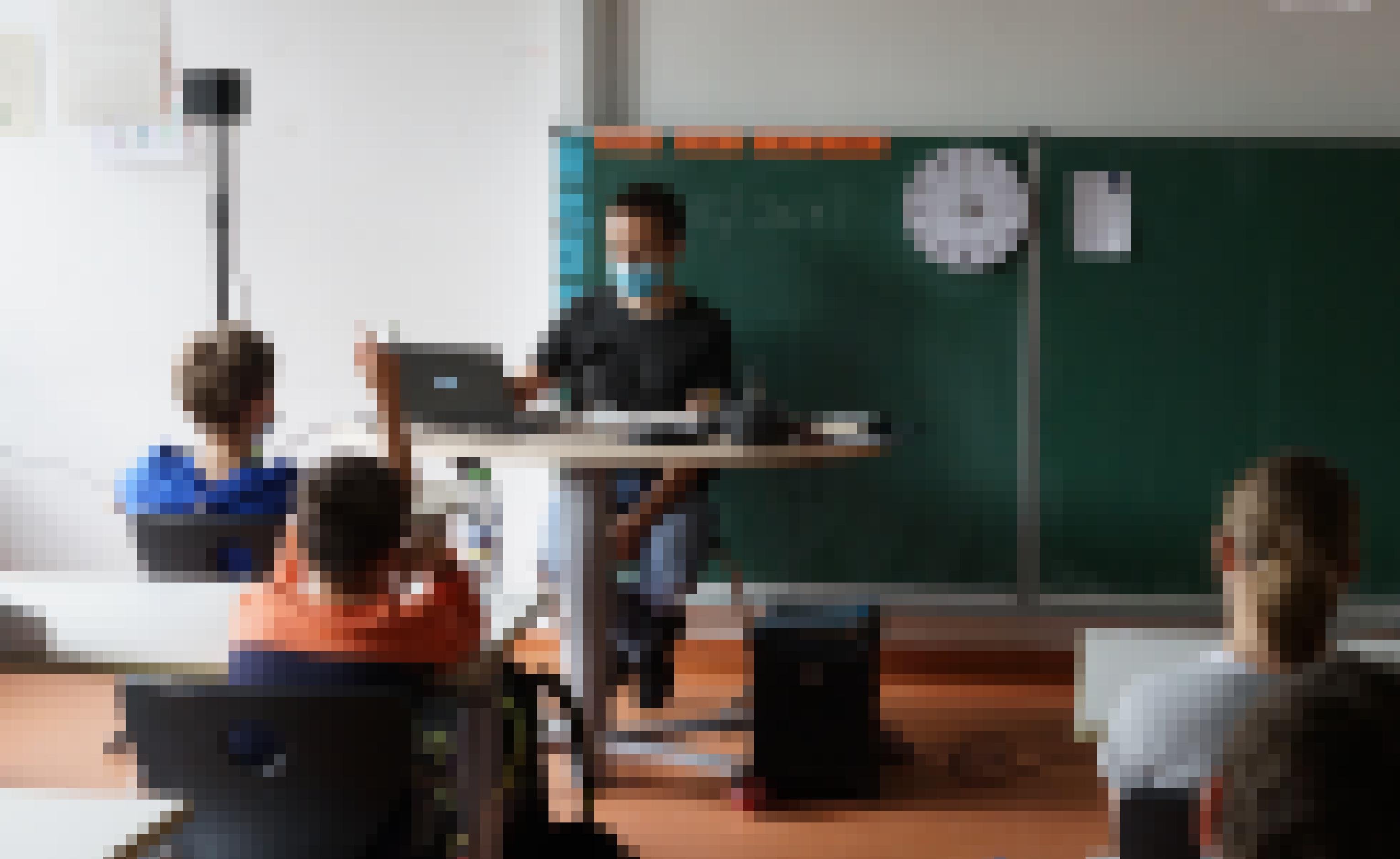 Ein junger Mann sitzt an einem Lehrertisch, mit Boxen, Laptop. Davor sitzen mehrere Schüler, einer meldet sich.