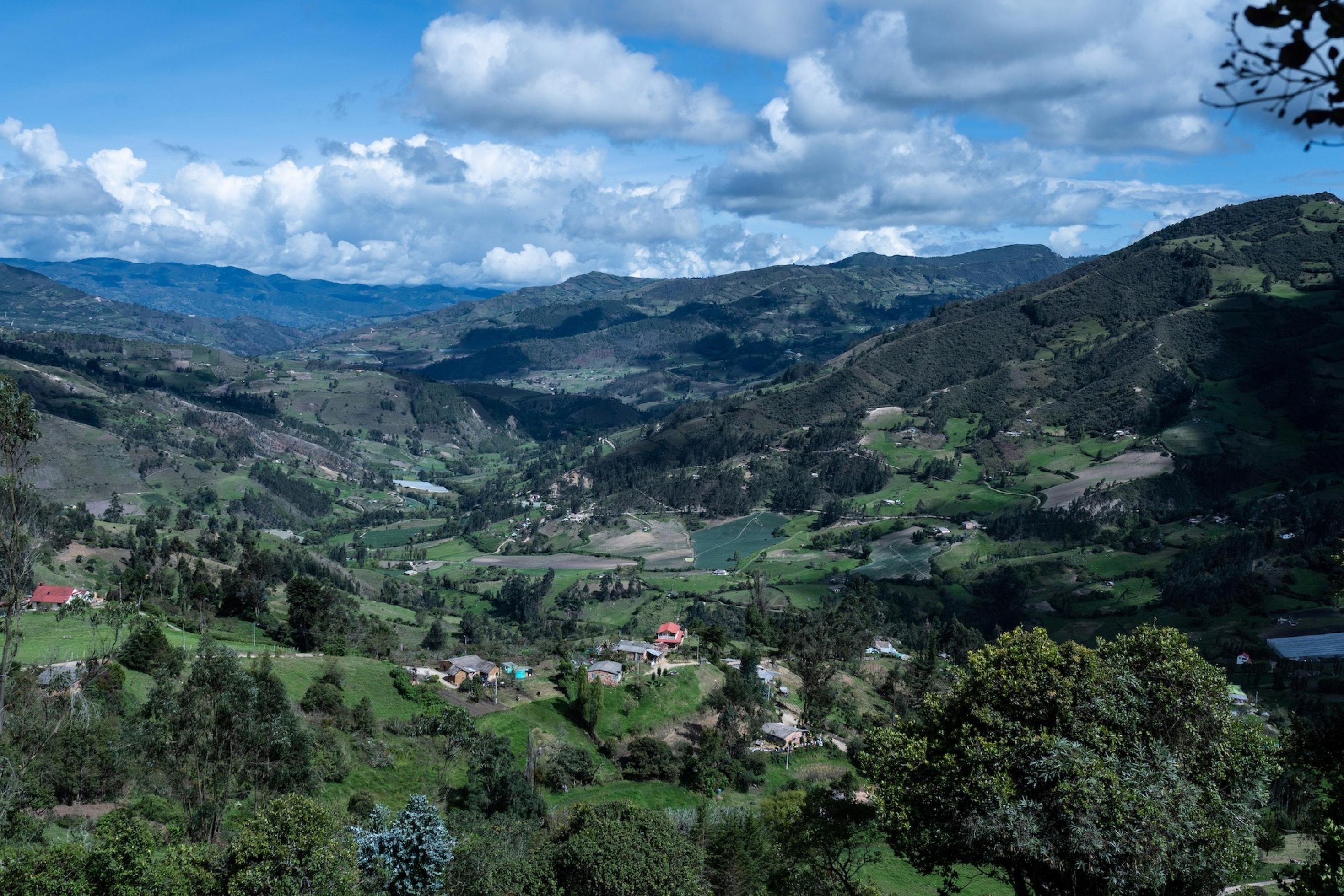 Blauer Himmel mit Wolken über bergiger, grüner Landschaft. Vereinzelt Häuser. Ein Flickenteppich aus kleinen Feldern, Ackern, Bäumen. Kaum mehr intakte Bewaldung auf den Bergkuppen.