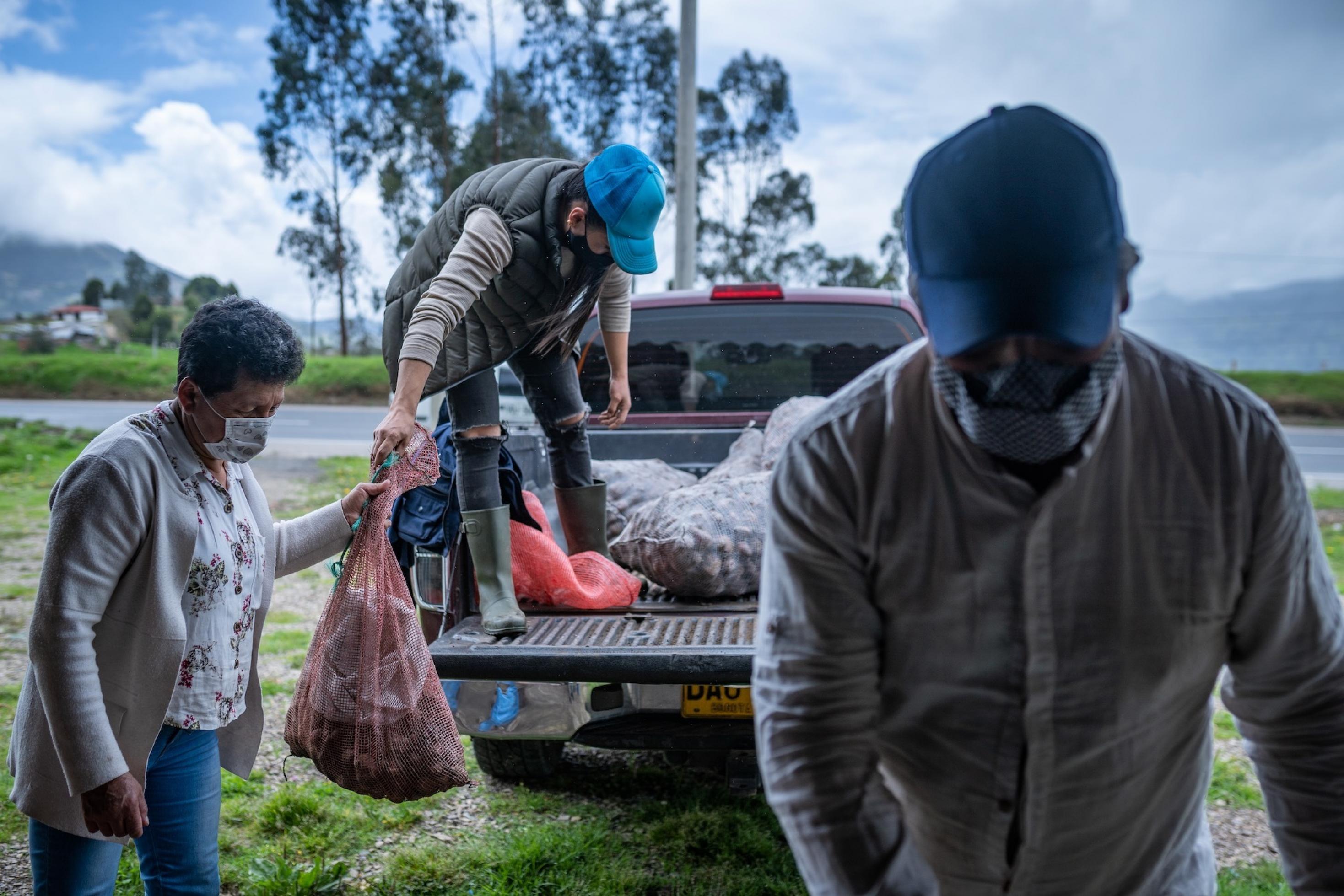 Tochter steht auf Ladefläche von Pick-Up, auf dem schon einige Kartoffelsäcke liegen. Mutter reicht ihr durchsichtigen Netz-Sacke mit mehreren Beuteln darin. Im Vordergrund blickt Vater nach unten und packt (nicht im Bild) den nächsten Sack. Alle tragen Mundschutz.