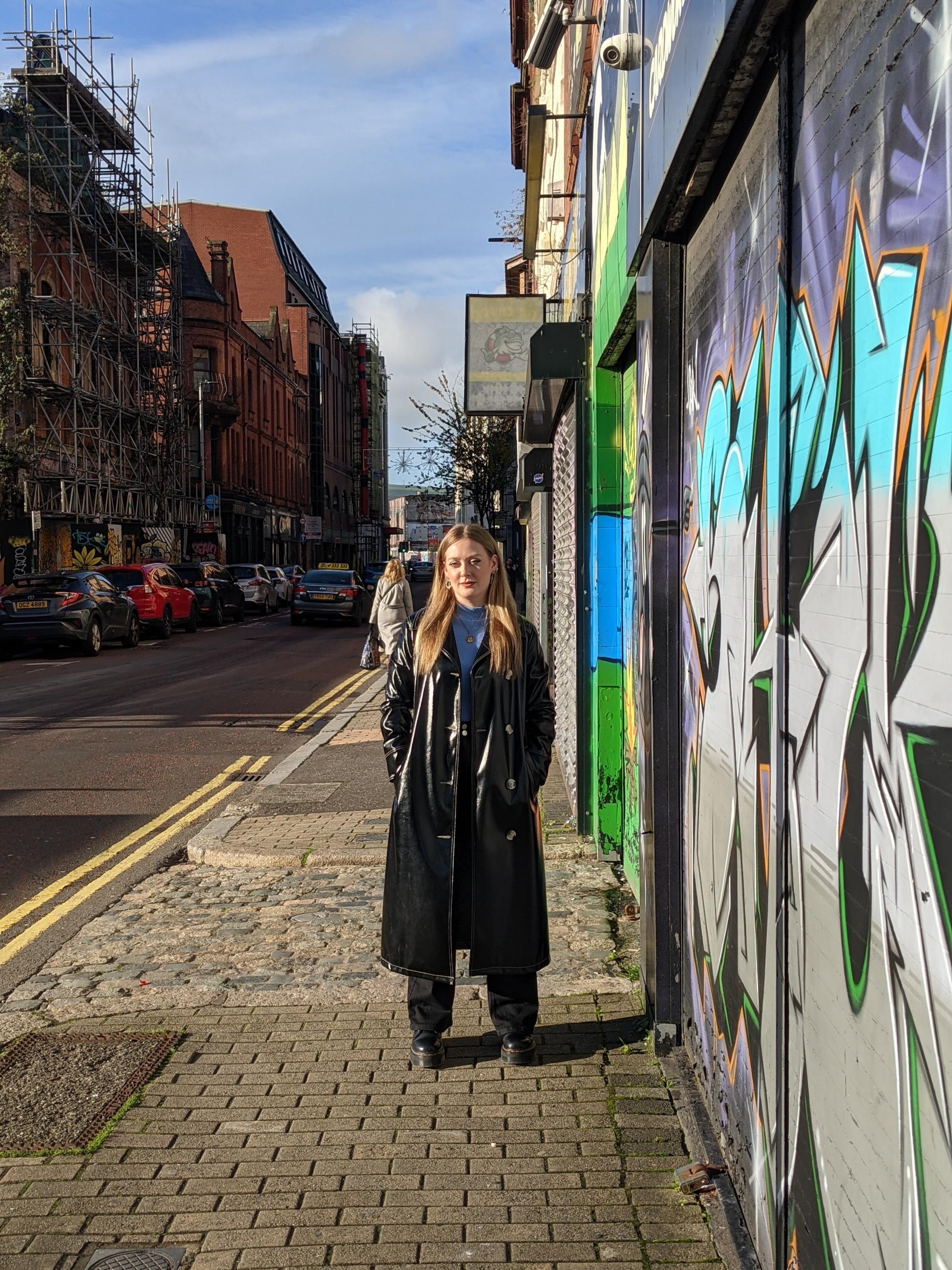 Eine junge Frau im schwarzen Mantel steht in einer Gasse in Belfast.