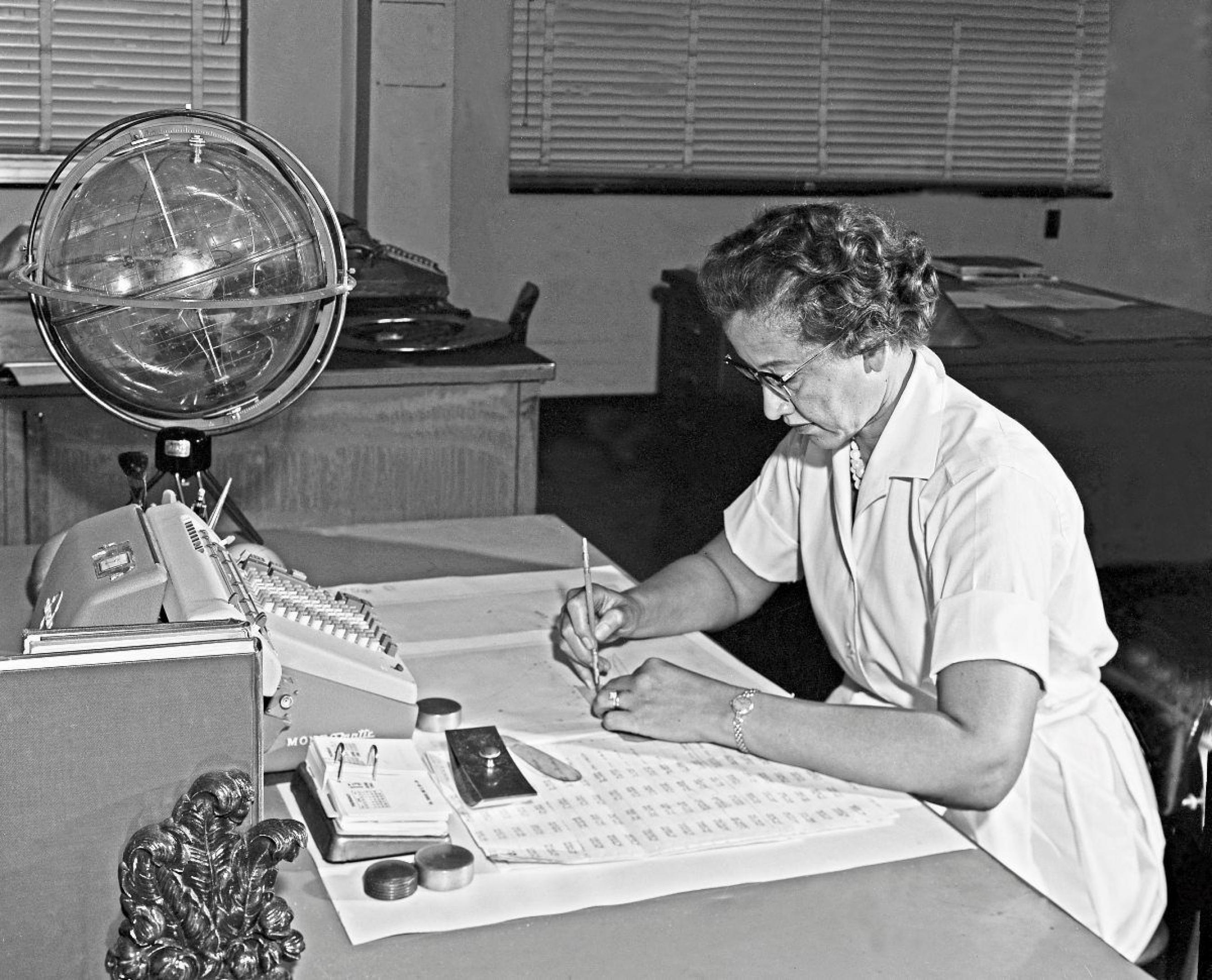 Die Mathematikerin Katherine Johnson sitzt an ihrem Schreibtisch und schreibt mit ihrem Rechenschieber und dem „Celestial Training Device“.