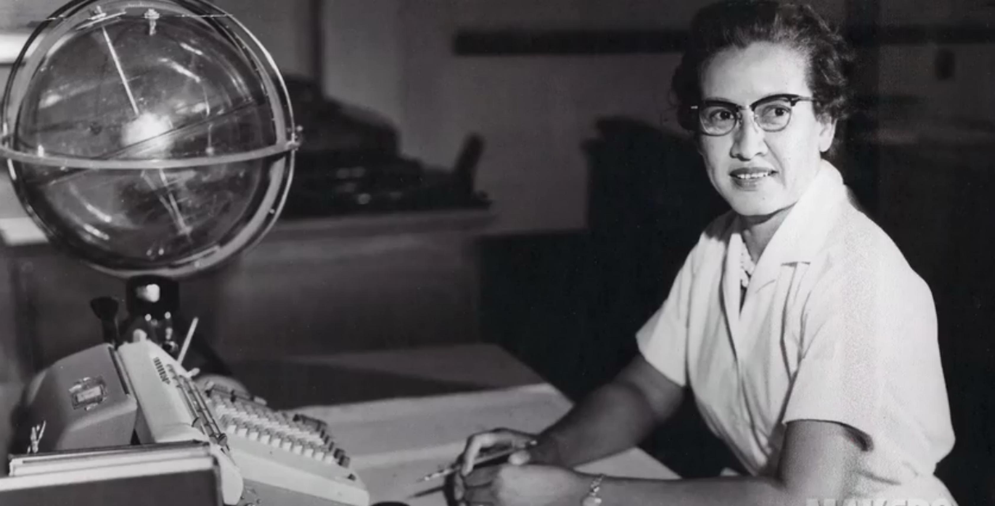 Rechts im Bild sitzt eine Frau am Schreibtisch; sie trägt einen weißen, kurzärmeligen Kittel und eine Brille; vor ihr auf dem Schreibtisch, im Bild links, steht eine Rechenmaschine und im Hintergrund ein Globus.