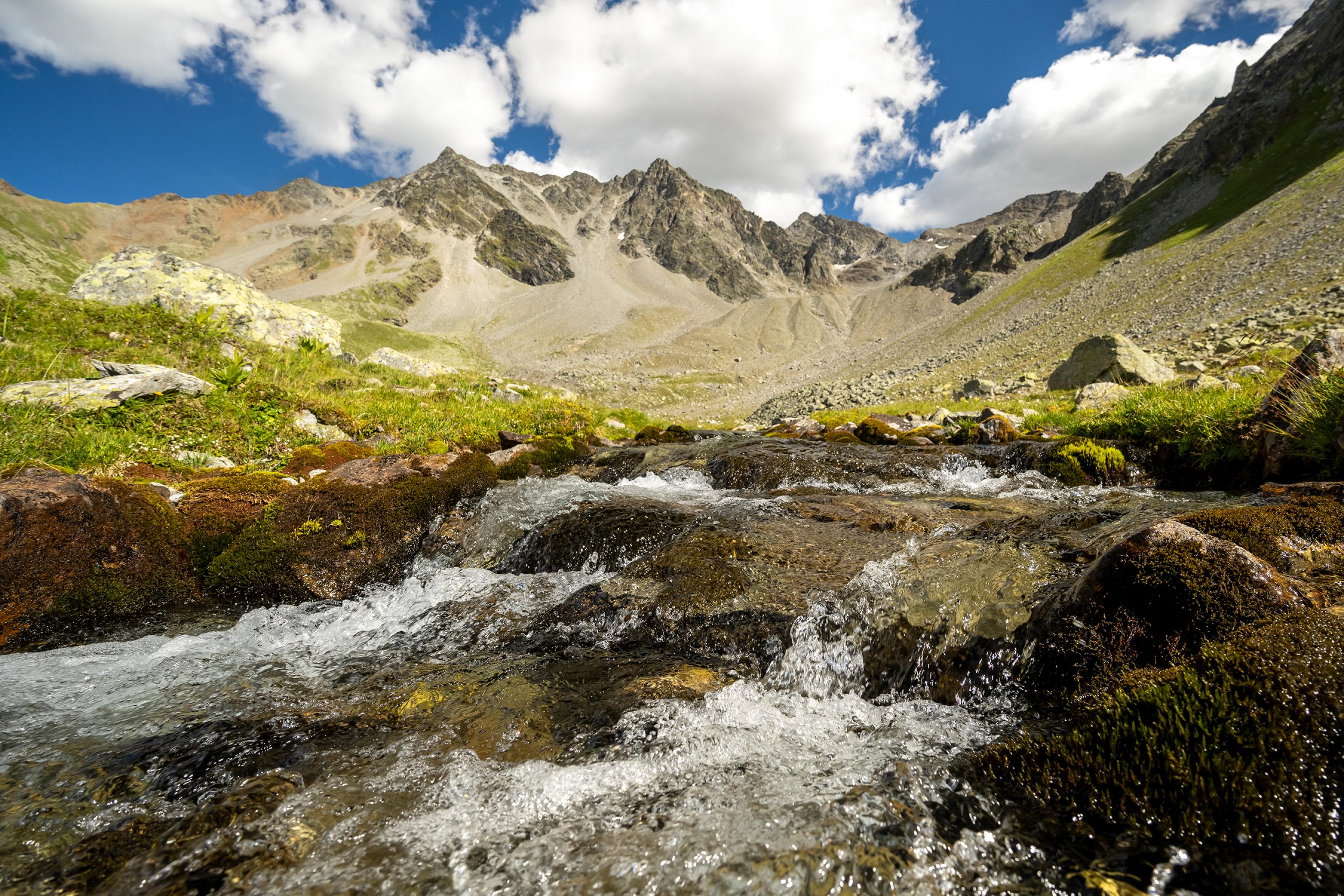 Gebirgsbach im Vordergrund, dahinter alpines Hochtal mit Almwiesen und Bergen