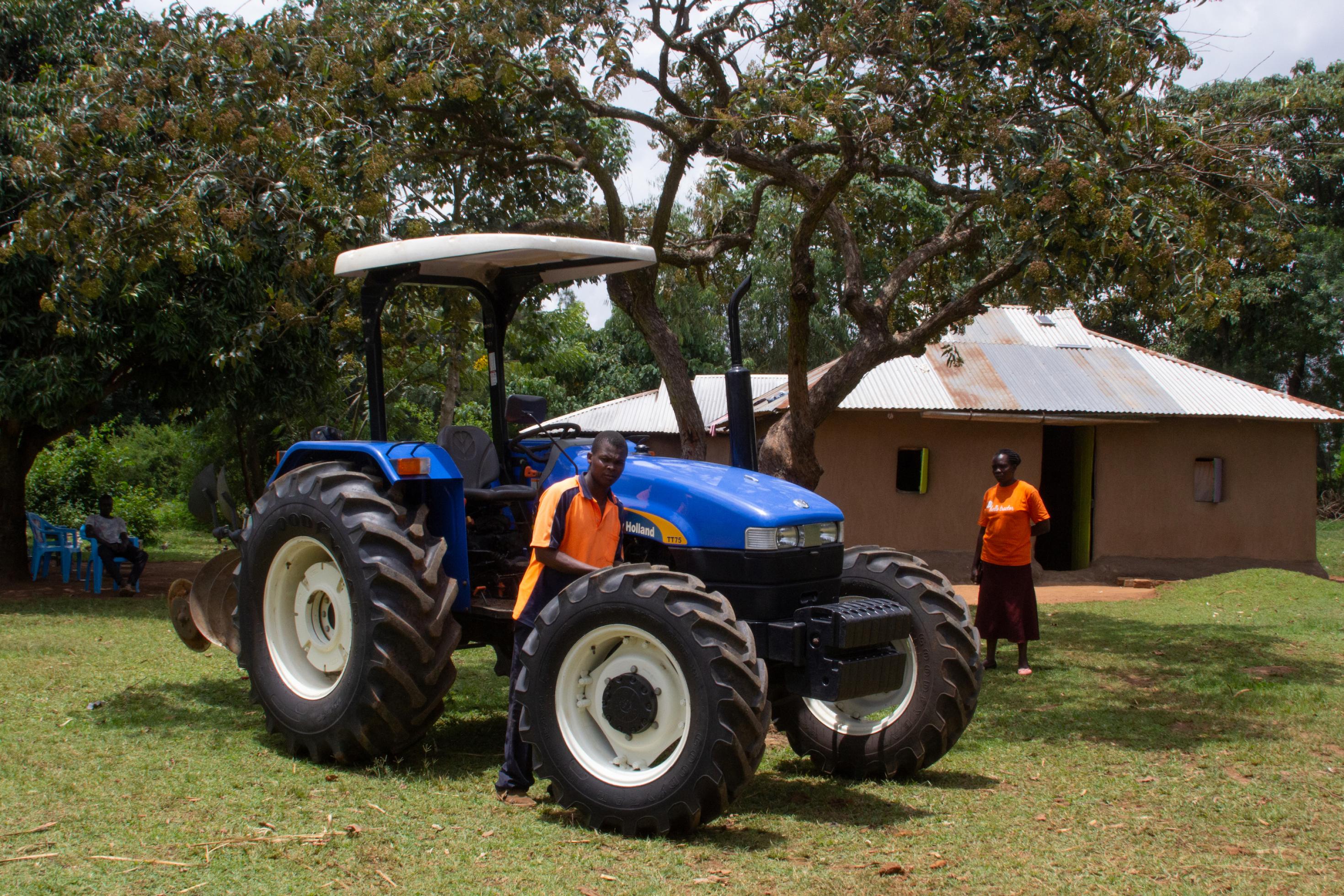 Der Traktor steht in der Bildmitte, rechts und links zwei Menschen, ein Haus im Hintergrund.