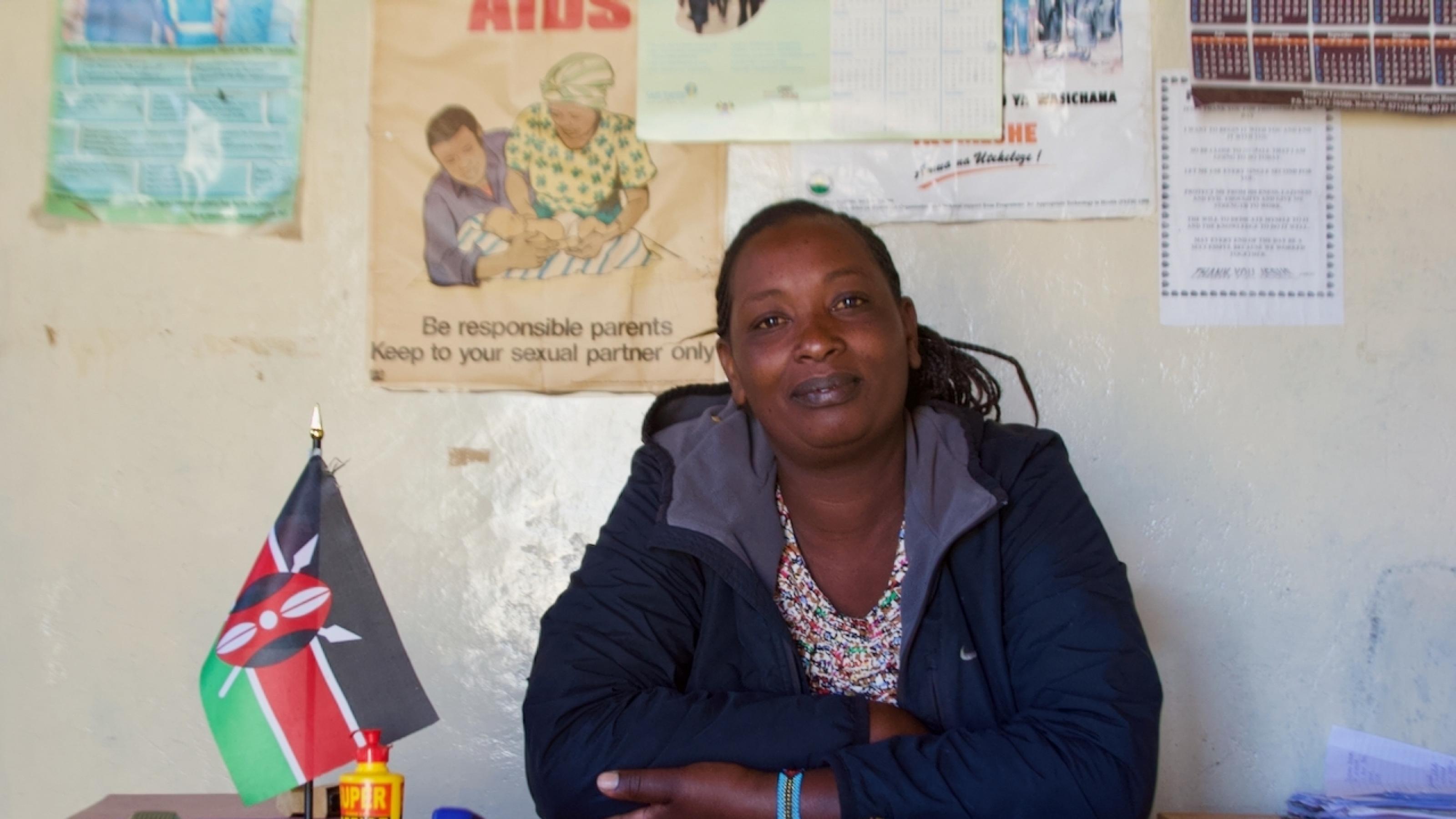 Eine Frau Ende 30 sitzt hinter einem einfachen Schreibtisch, darauf eine kenianische Standflagge, ein Fläschchen mit Tipp-Ex, einige Dokumente. Sie guckt selbstbewusst in die Kamera. An der Wand einige Poster zur Aufklärung über Frauenrechte und Ähnliches.