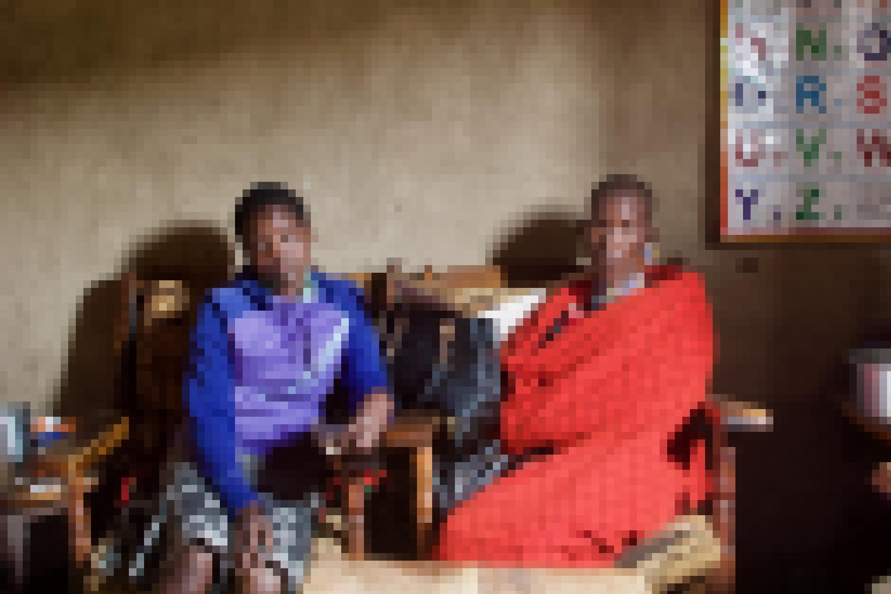 Mutter und Tochter sitzen in zwei Sesseln nebeneinander. Die Mutter hat sich in ein typisches, rotes Massaituch gewickelt, außerdem trägt sie Massai-Schmuck. Die Tochter trägt einen warmen Kapuzenpulli und einen Rock.  Im Hintergrund sind die Lehmwände des Hauses zu sehen, und im Anschnitt ein Wandposter mit großen Buchstaben zum Lesen-Lernen.