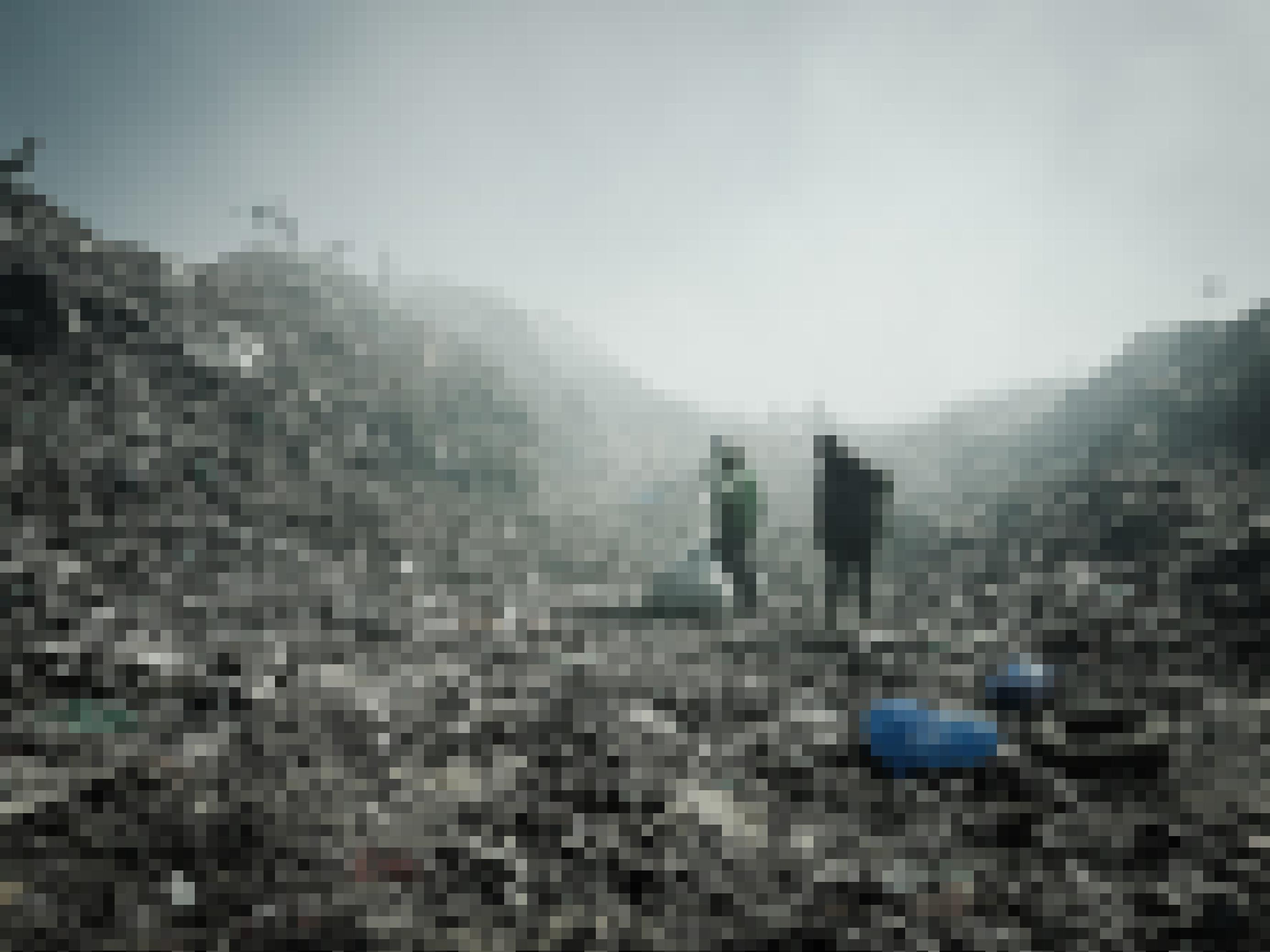Zwischen hohen Bergen aus Müll zwei Menschen, einer trägt einen Sack, der andere schleift einen Sack hinterher. Die Luft ist von Rauch geschwängert. Auf der Spitze der Müllberge links im Bild sitzen einige Marabout genannte Vögel, es sind Aasfresser.