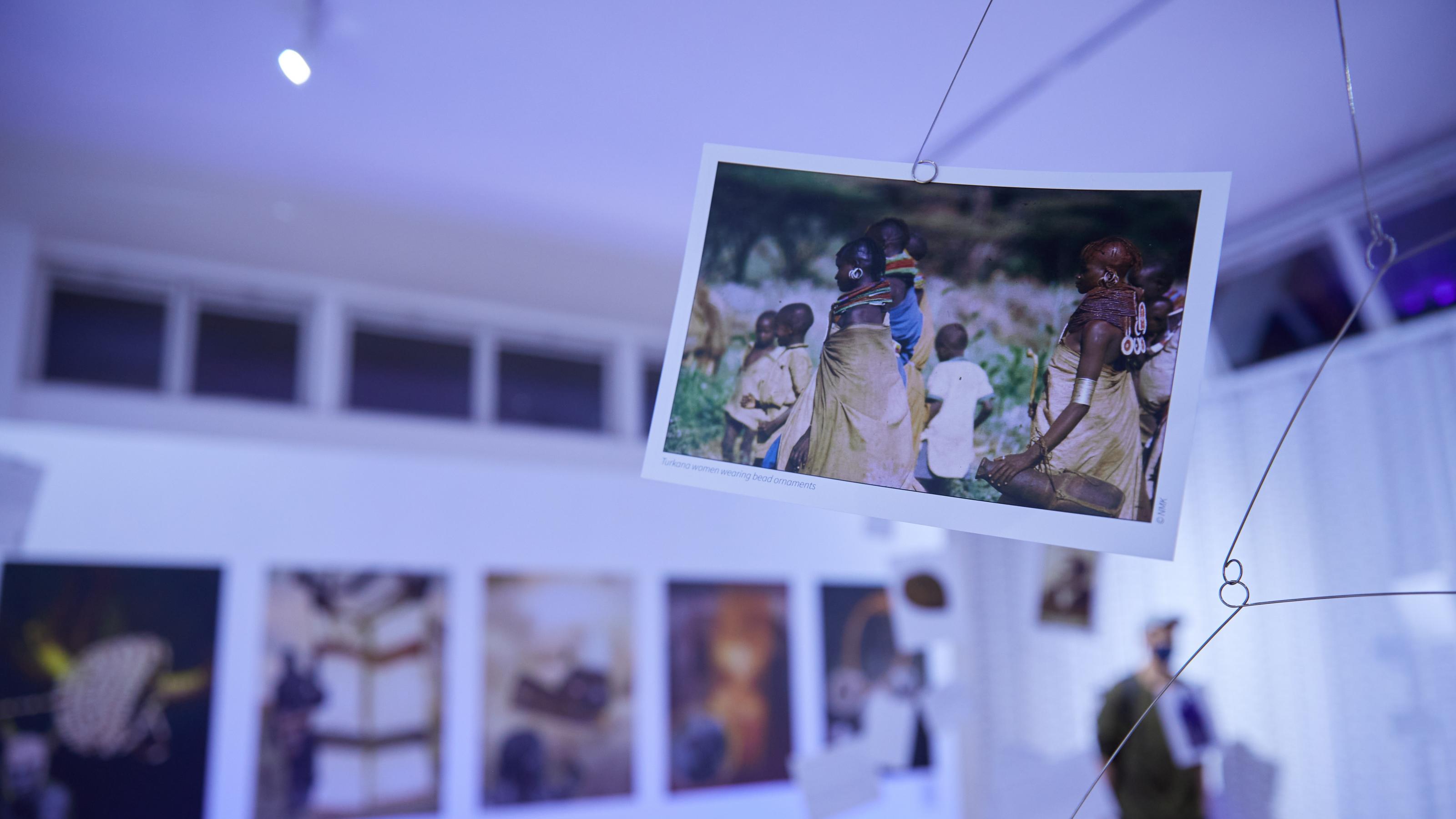Im Mittelgrund hängt ein Foto an einer Art Mobile, darauf sind traditionell gekleidete Kenianerinnen und Kenianer zu sehen, die auffällige Schmuckstücke tragen. Im Hintergrund und verschwommen weitere Fotos.