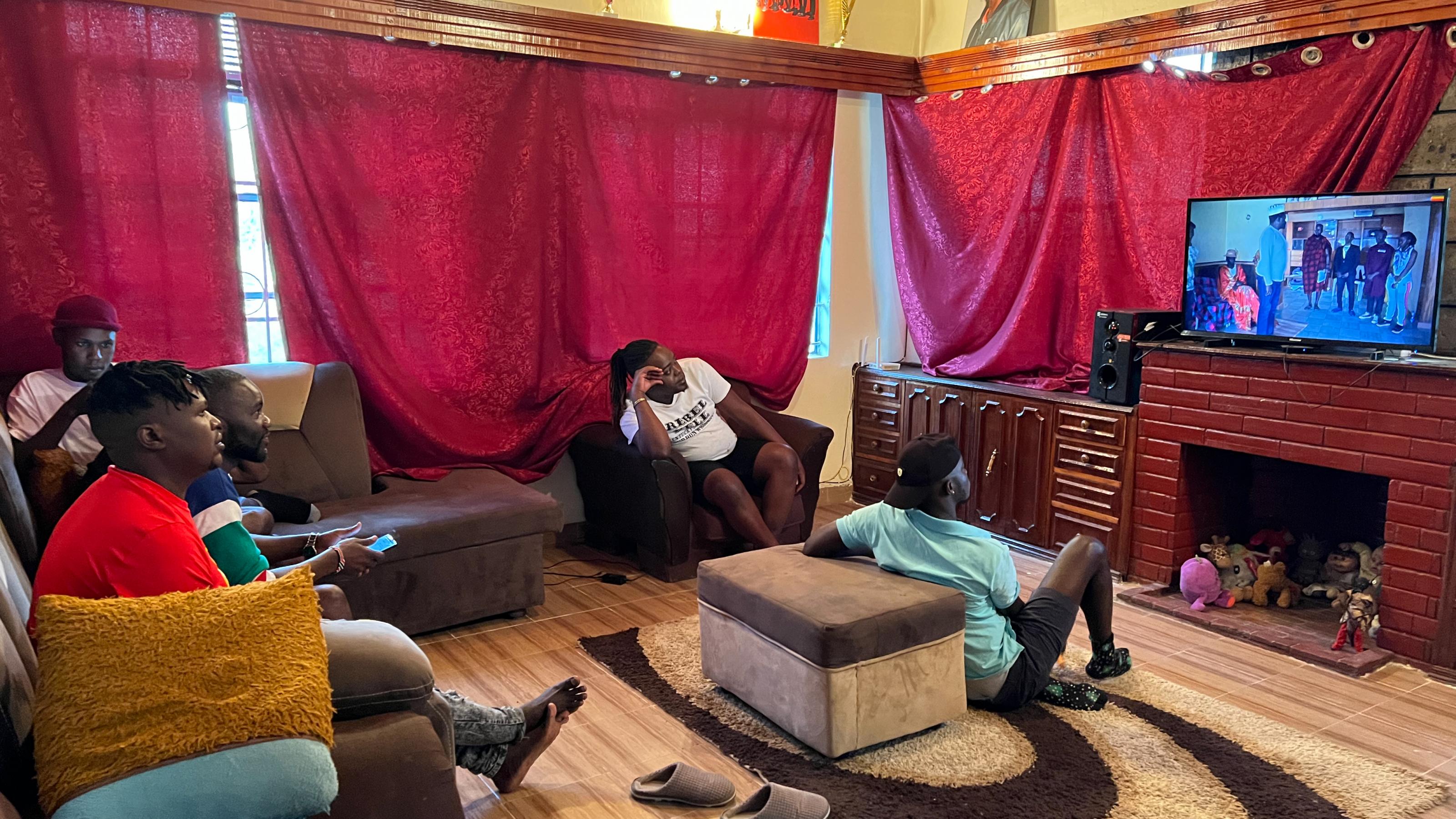 Zu sehen sind einige Männer, die in einem Wohnzimmer gemeinsam Fernsehen.