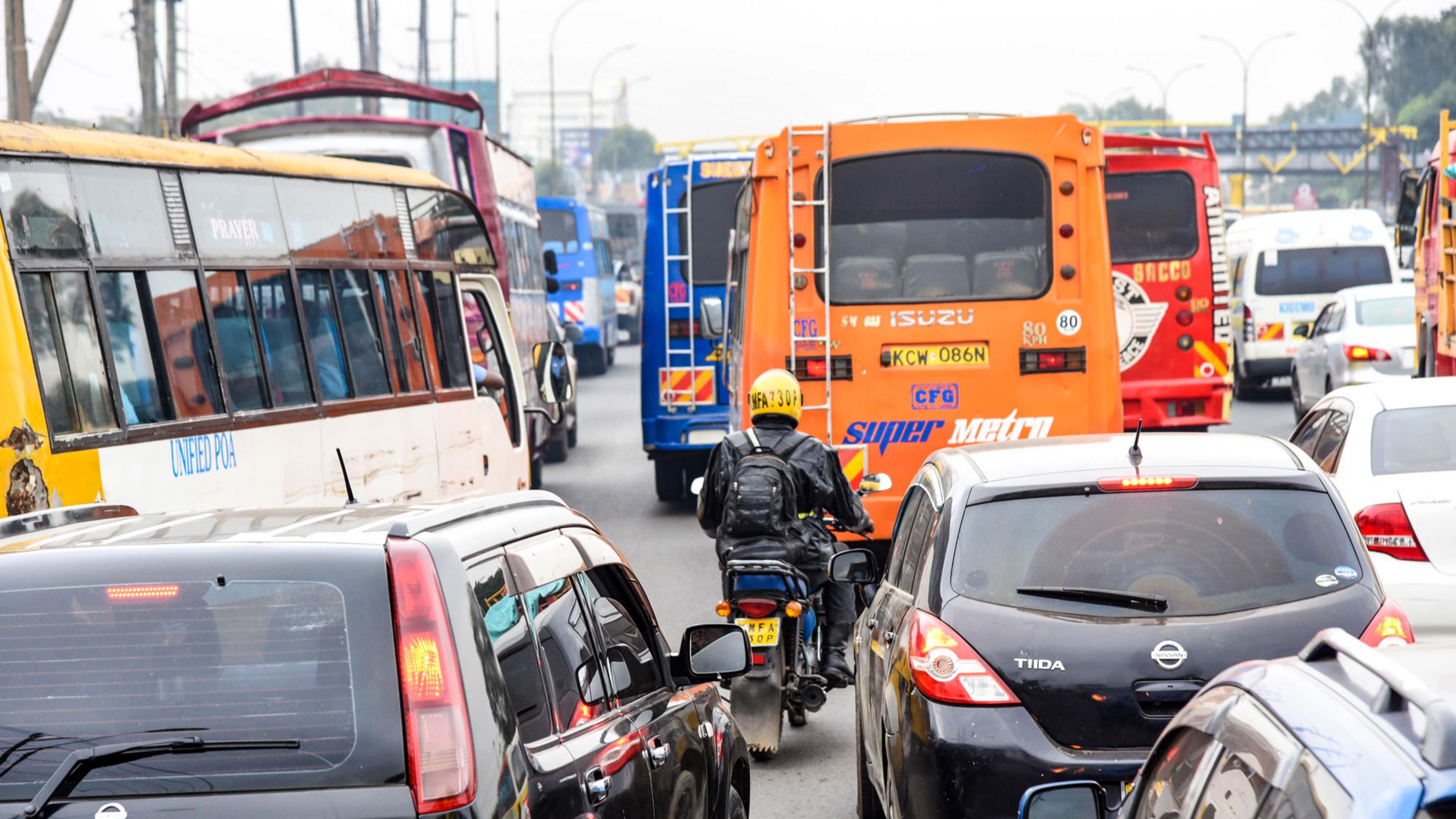Das Foto zeigt dichten Verkehr und einen Motorradfahrer von hinten, der durch Schlangen stehender Fahrzeuge hindurch fährt.