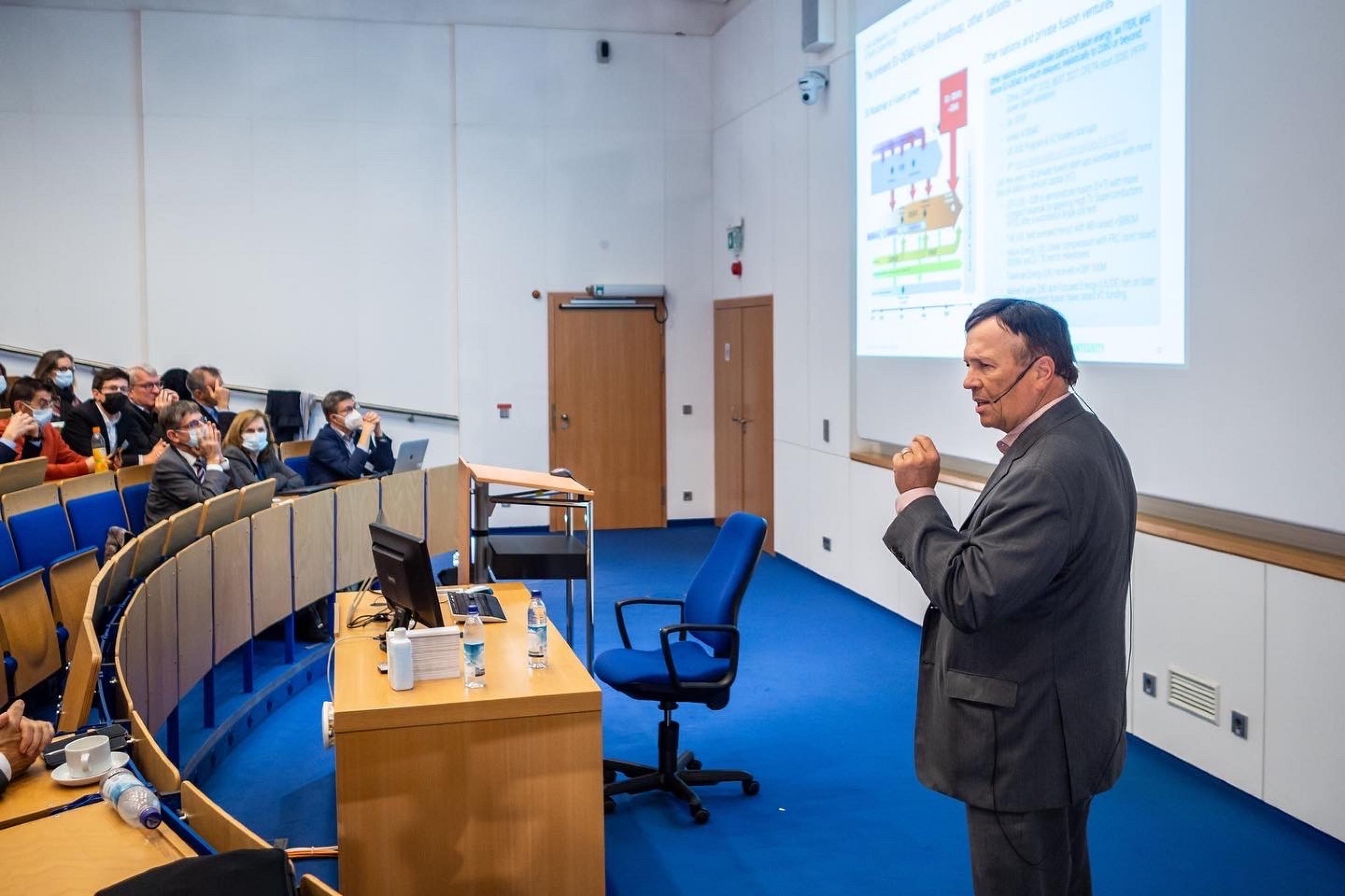 Ein Mann hält vor Publikum eine Power Point Präsentation und hat ein Mikrofon in der Hand.