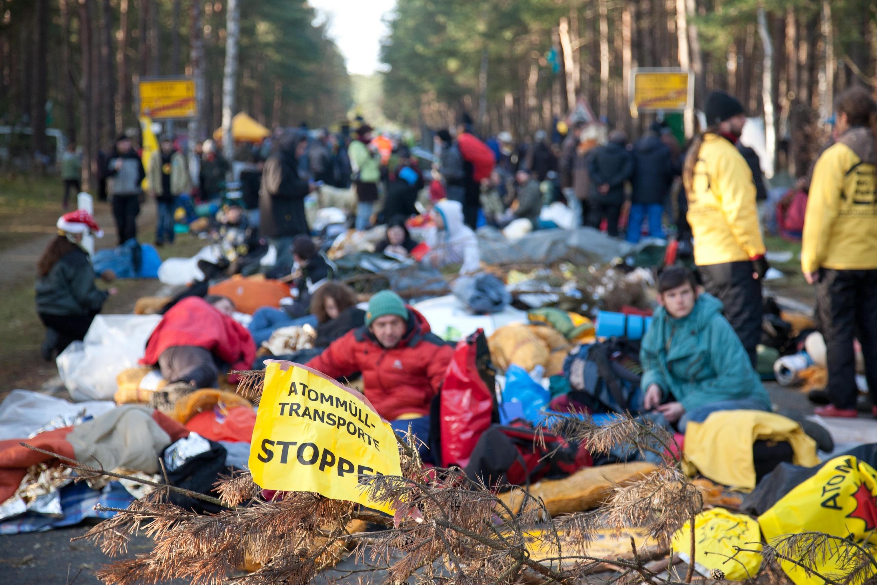 Menschen sitzen mit Schildern gegen Atomkraft auf einer Straße in Richtung Gorleben und blockieren sie. Auf einem Schild steht „Atomtransport stoppen“