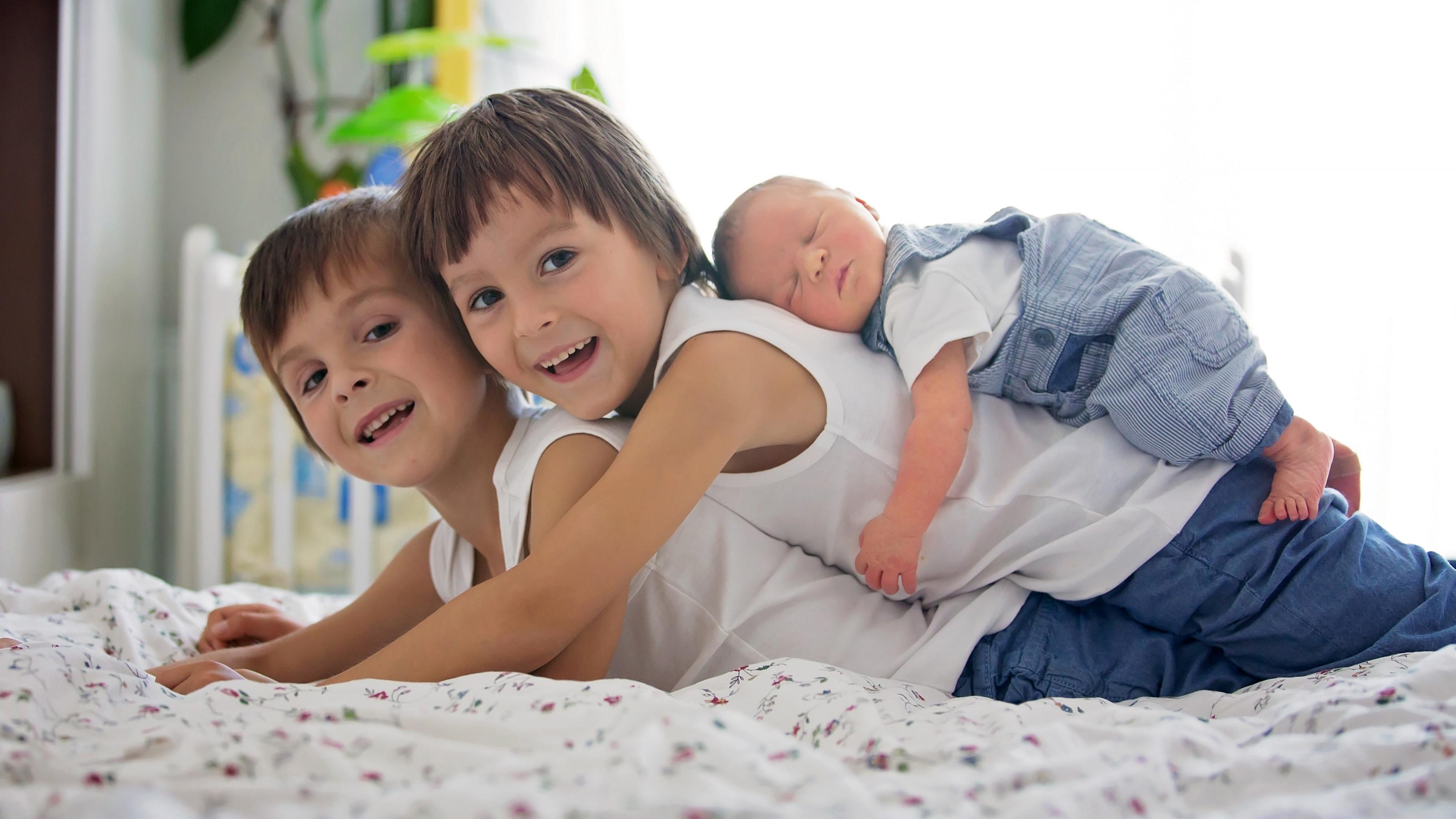 Zwei kleine Jungen liegen auf einem Bett. Auf dem Rücken des einen liegt ein neugeborenes Baby mit geschlossenen Augen.