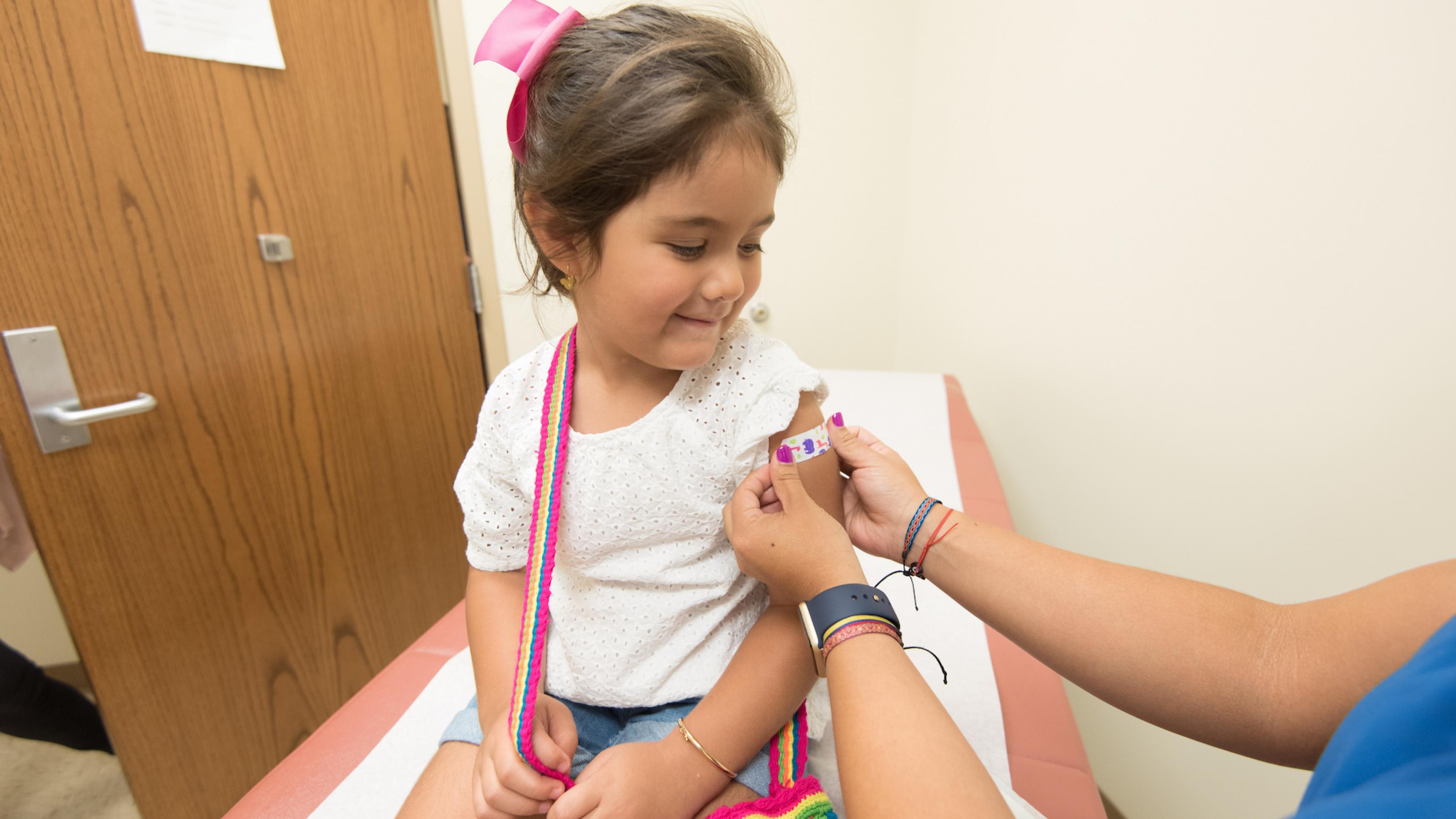 Auf dem Bild ist ein kleines Mädchen mit brauchen Haaren und einer bunten Umhängetasche zu sehen, die gerade ein Pflaster auf den Oberarm bekommt, weil sie bei der Ärztin eine Impfung erhalten hat.