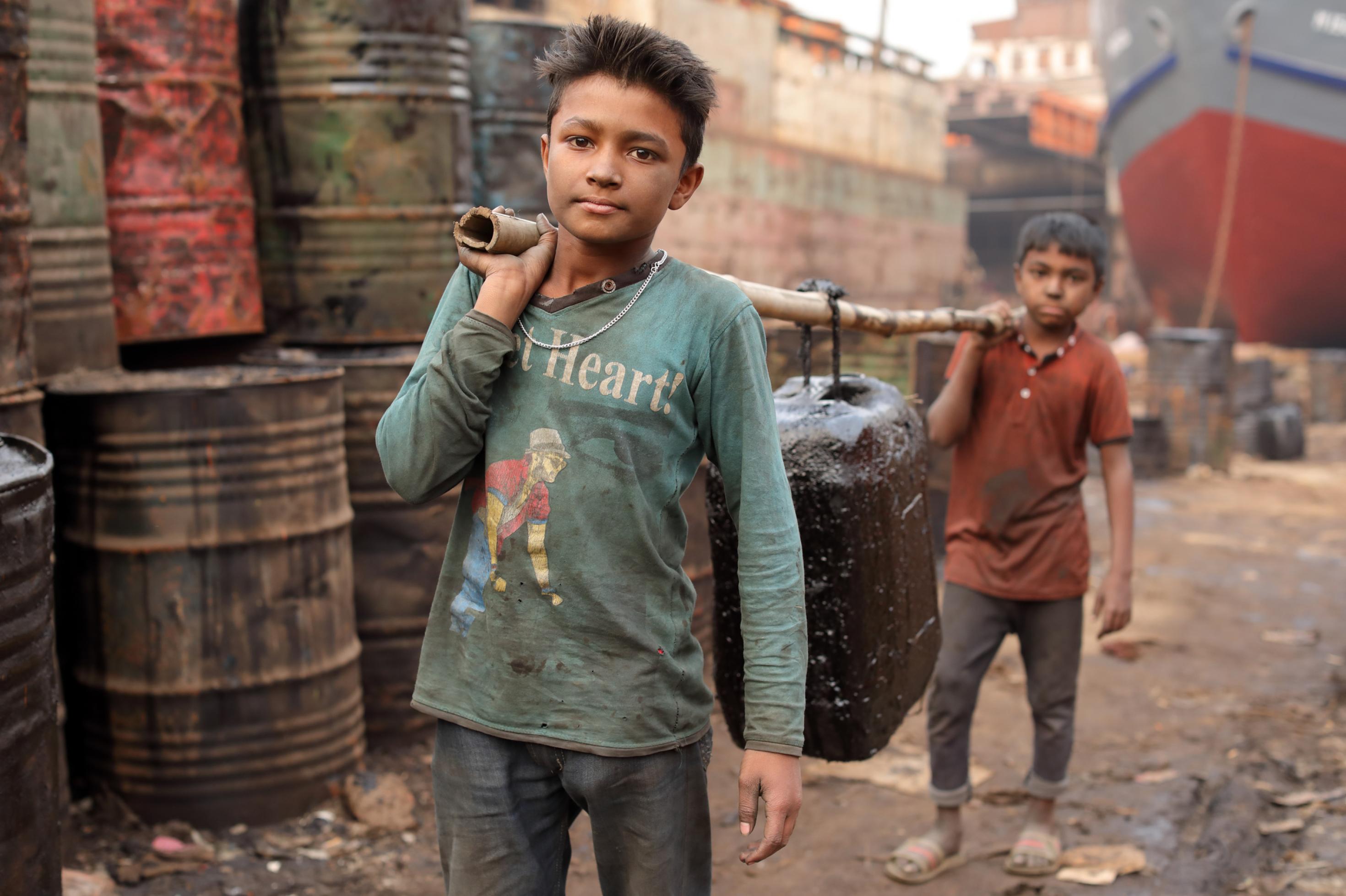 Das Foto zeigt eine Werft, links im Bild sind Fässer aufgestapelt. Im Fokus des Bildes sind zwei etwa 10-jährige Jungen. Die beiden Kinderarbeiter tragen einer Stange, an der ein Kanister hängt. Mehr als 7,4 Millionen Mädchen und Jungen arbeiten, um nicht zu verhungern.