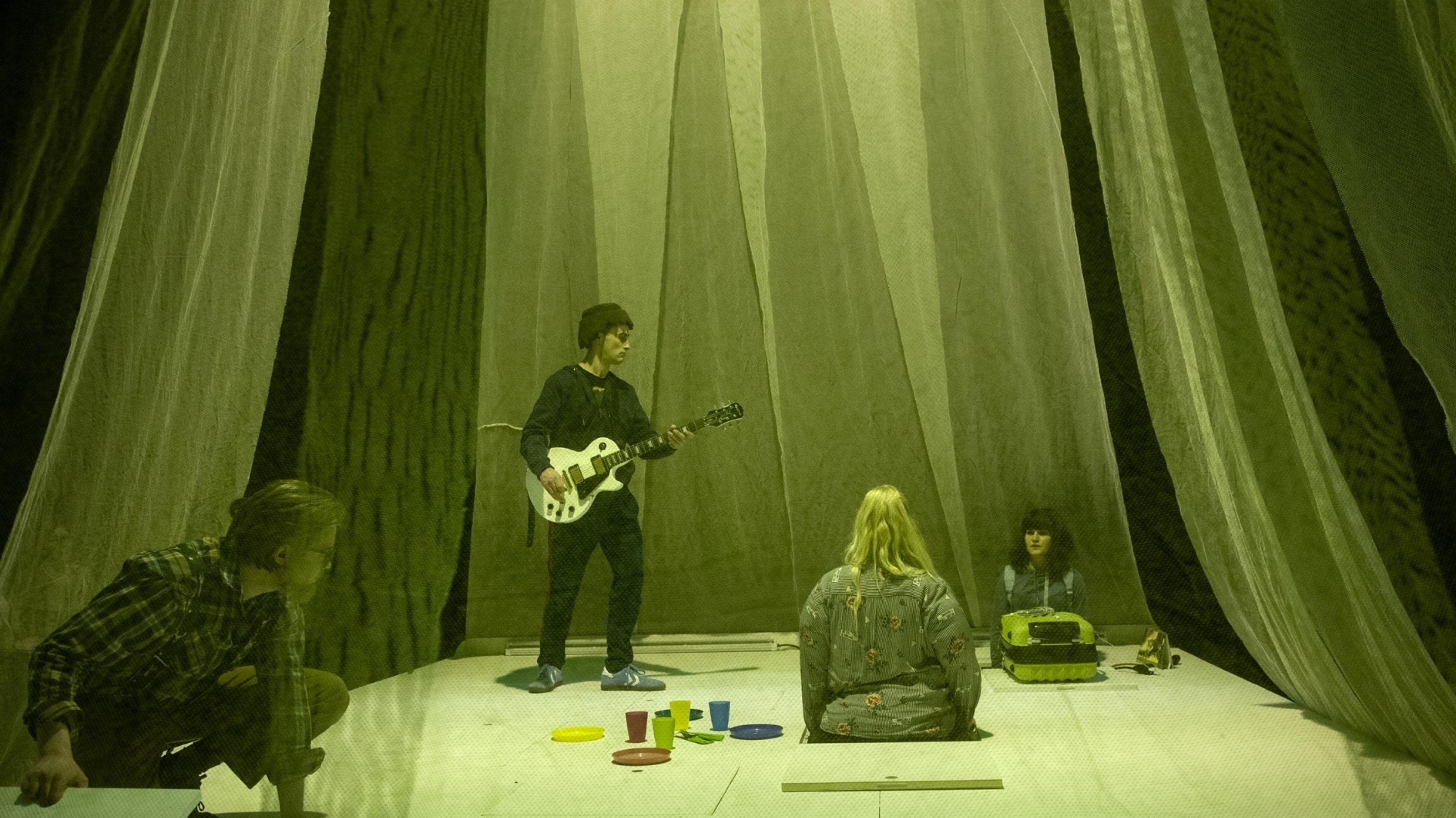 Zwei Darsteller und zwei Darstellerinnen spielen auf einer kleinen weißen Bühne, deren Bodenplatten man heben kann. Sie ist umgeben von transparenten Vorhängen. Das Licht ist grün.