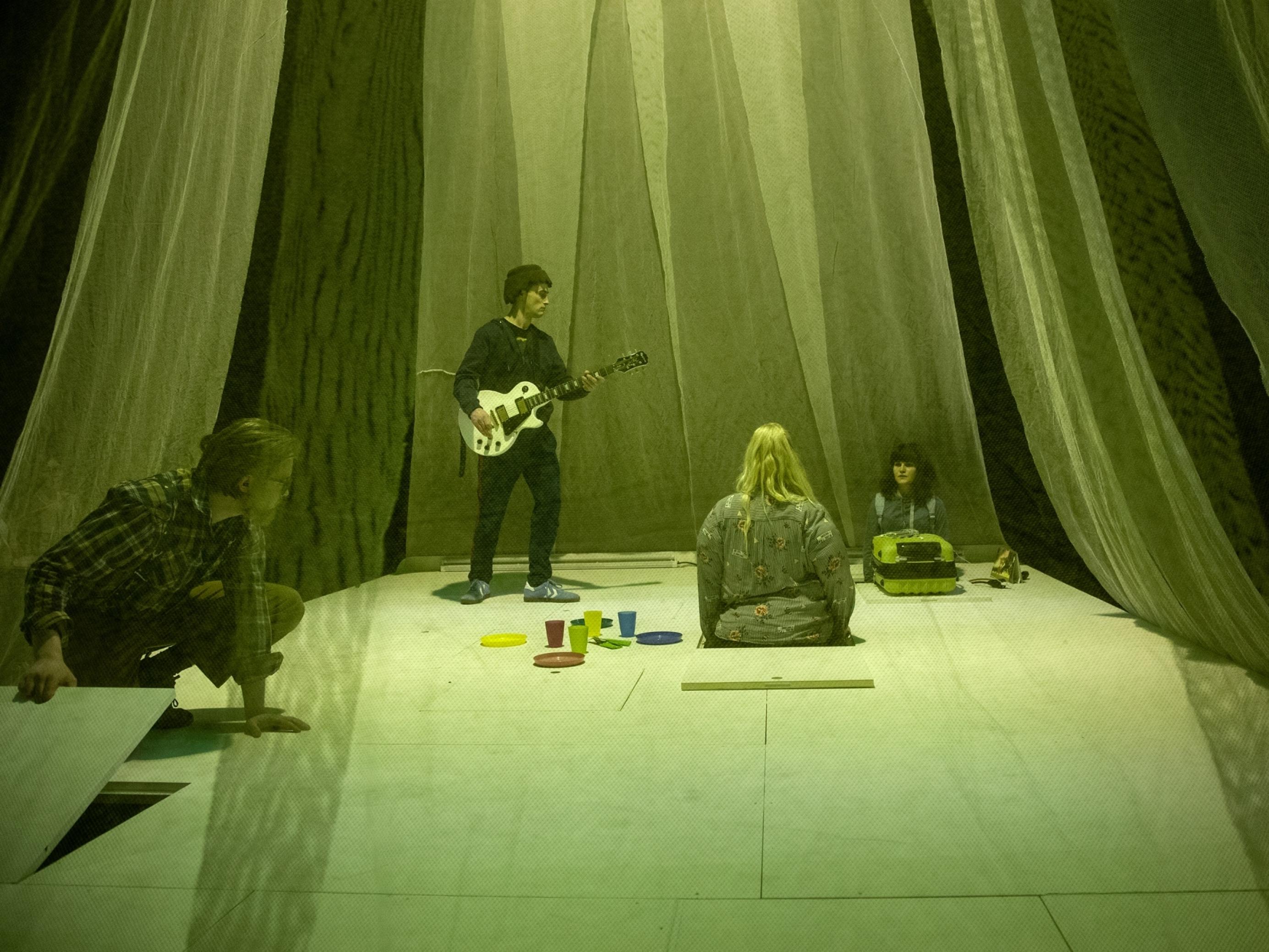 Zwei Darsteller und zwei Darstellerinnen spielen auf einer kleinen weißen Bühne, deren Bodenplatten man heben kann. Sie ist umgeben von transparenten Vorhängen. Das Licht ist grün.