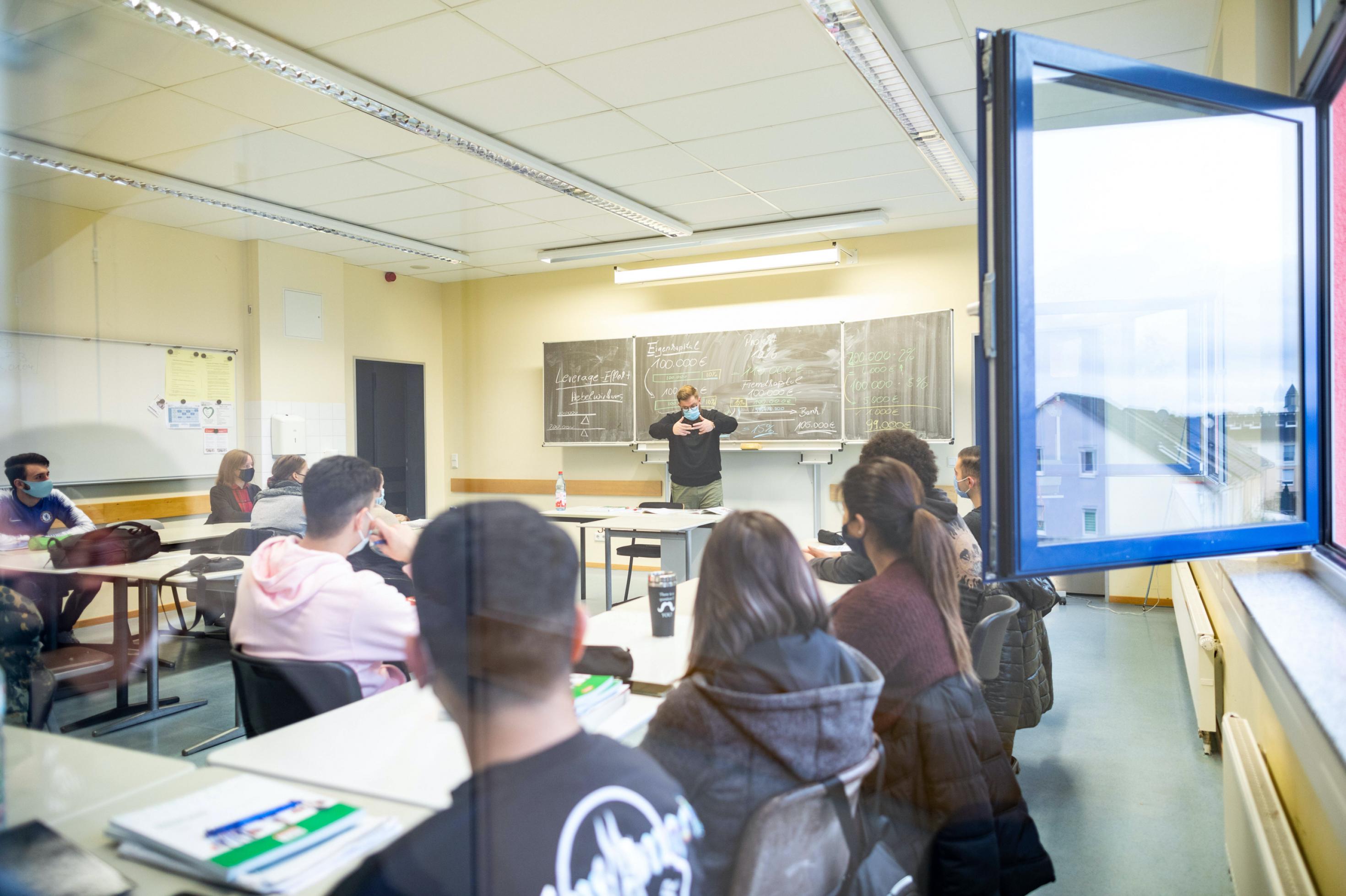 Teenager mit Jacken und dicken Pullis sitzen in einem Klassenraum, vorne an der Tafel steh der Lehrer. Alle tragen Gesichtsmasken. Die Fenster sind geöffnet.