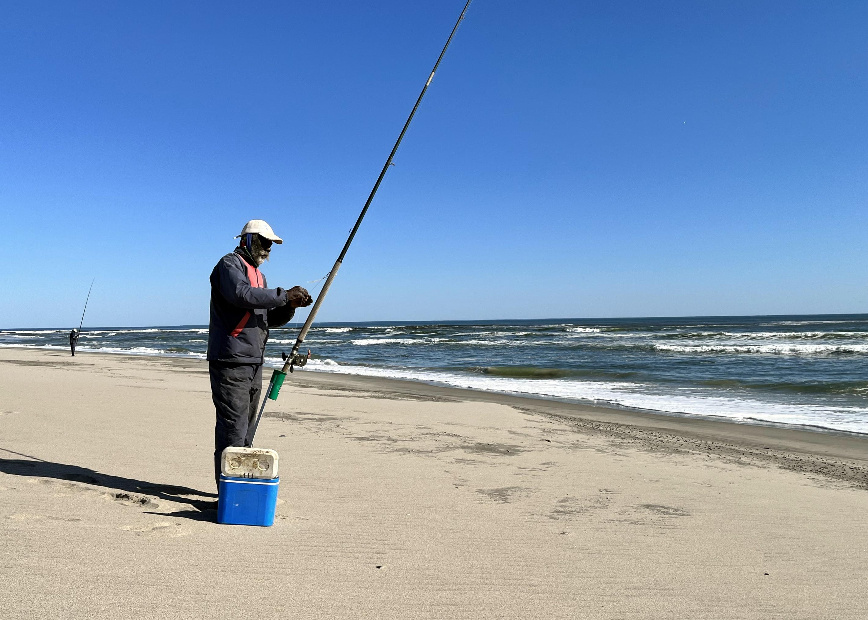 Der Fischer steht neben Angel und Kühlbox am Strand, er trägt eine blaue Jacke und hat einen grauen Bart.