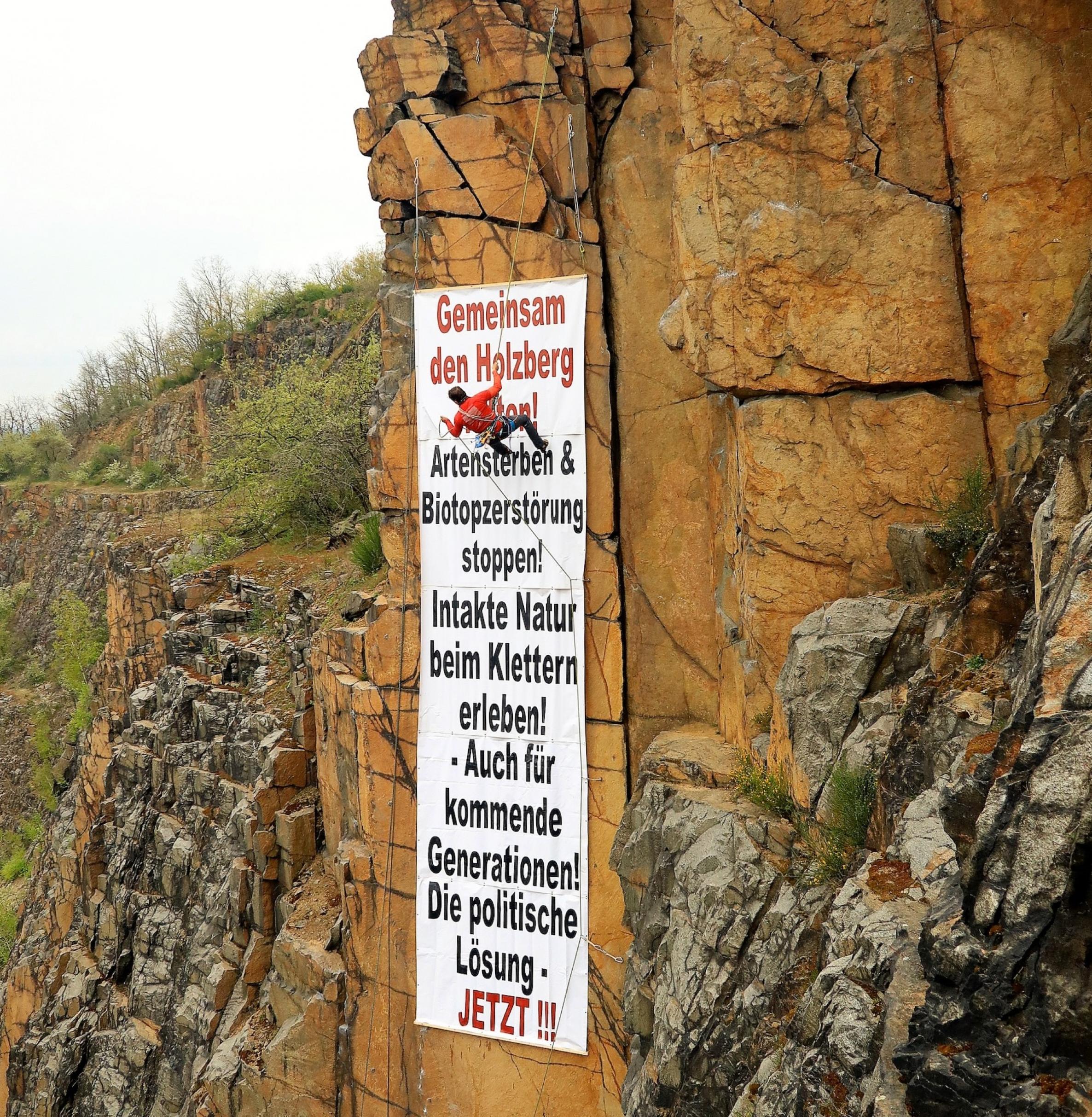 „Gemeinsam den Holzberg retten“ steht als Überschrift auf einem haushohen Plakat, das ein Kletterer an einer senkrechten Felswand anbringt.