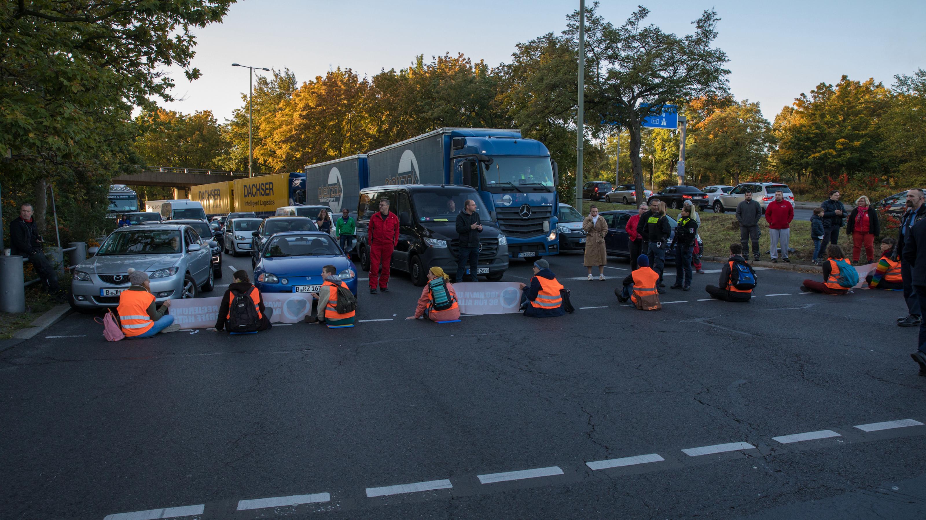 Menschen in orangefarbenen Warnwesten sitzen an einer Autobahnabfahrt auf der Straße und blockieren den Verkehr.