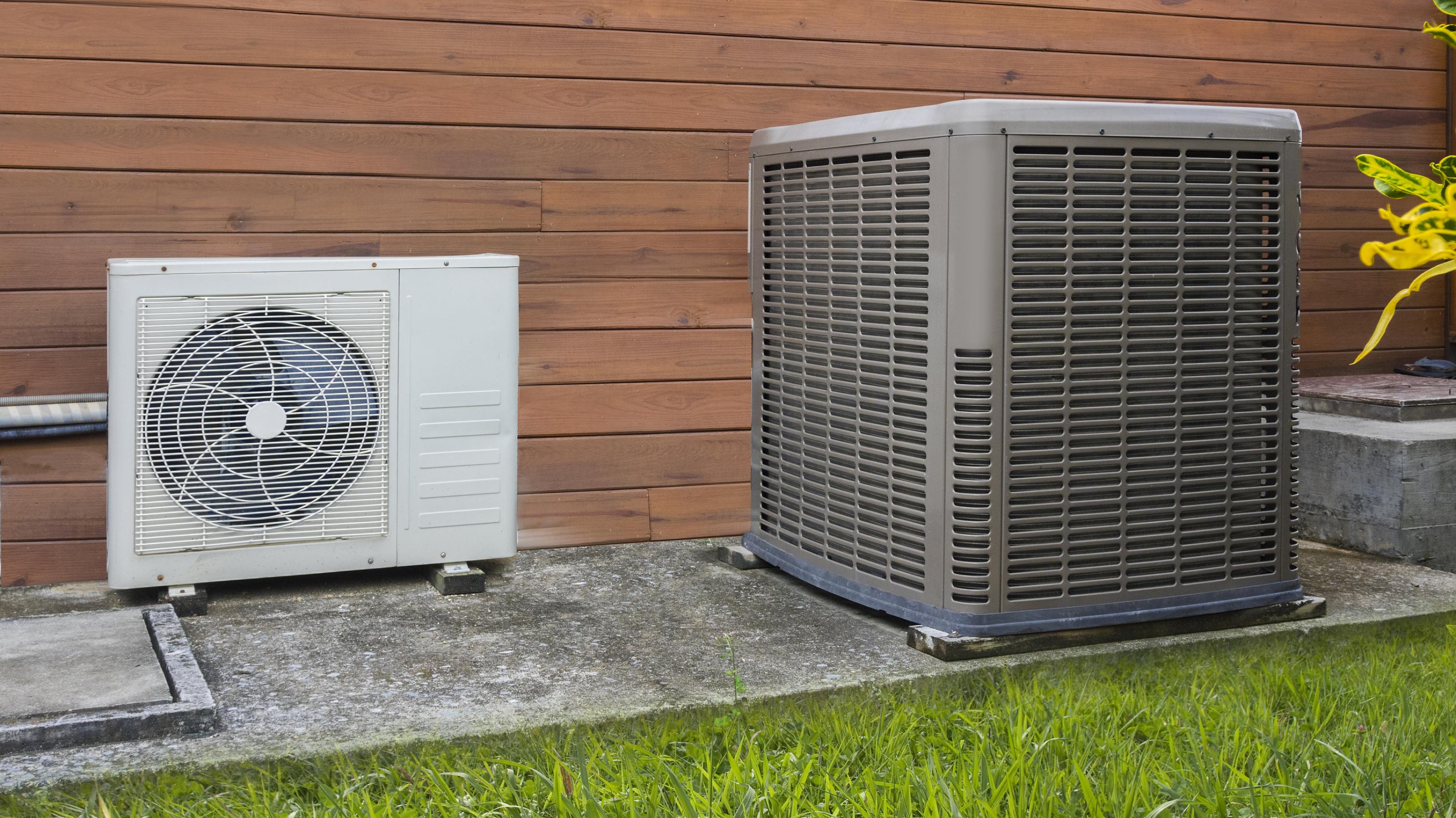 An der Außenseite eines Hauses stehen zwei unterschiedliche Klimaanlagen-Wärmepumpen auf einem Betonfundament.