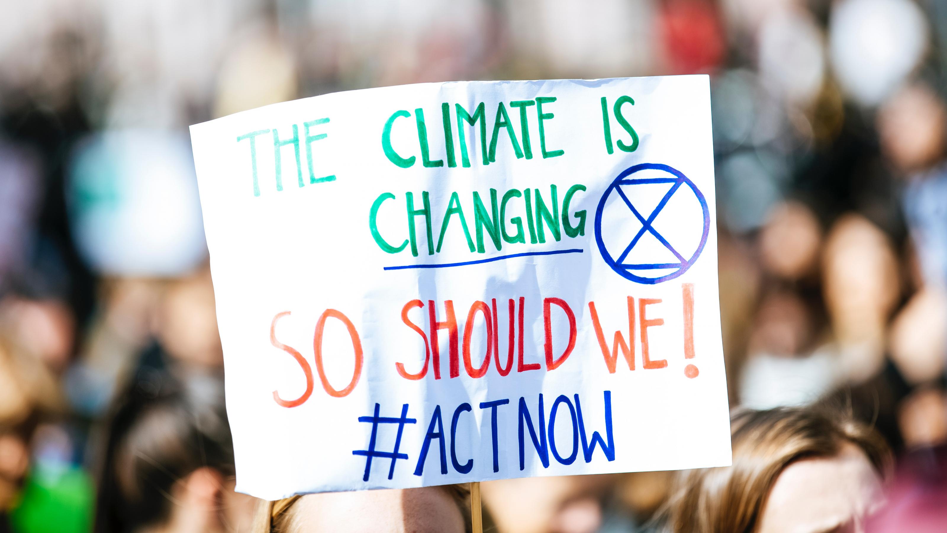 Demonstrierende halten ein Schild hoch auf dem steht: „The Climate is changing. So should we! #ActNow“