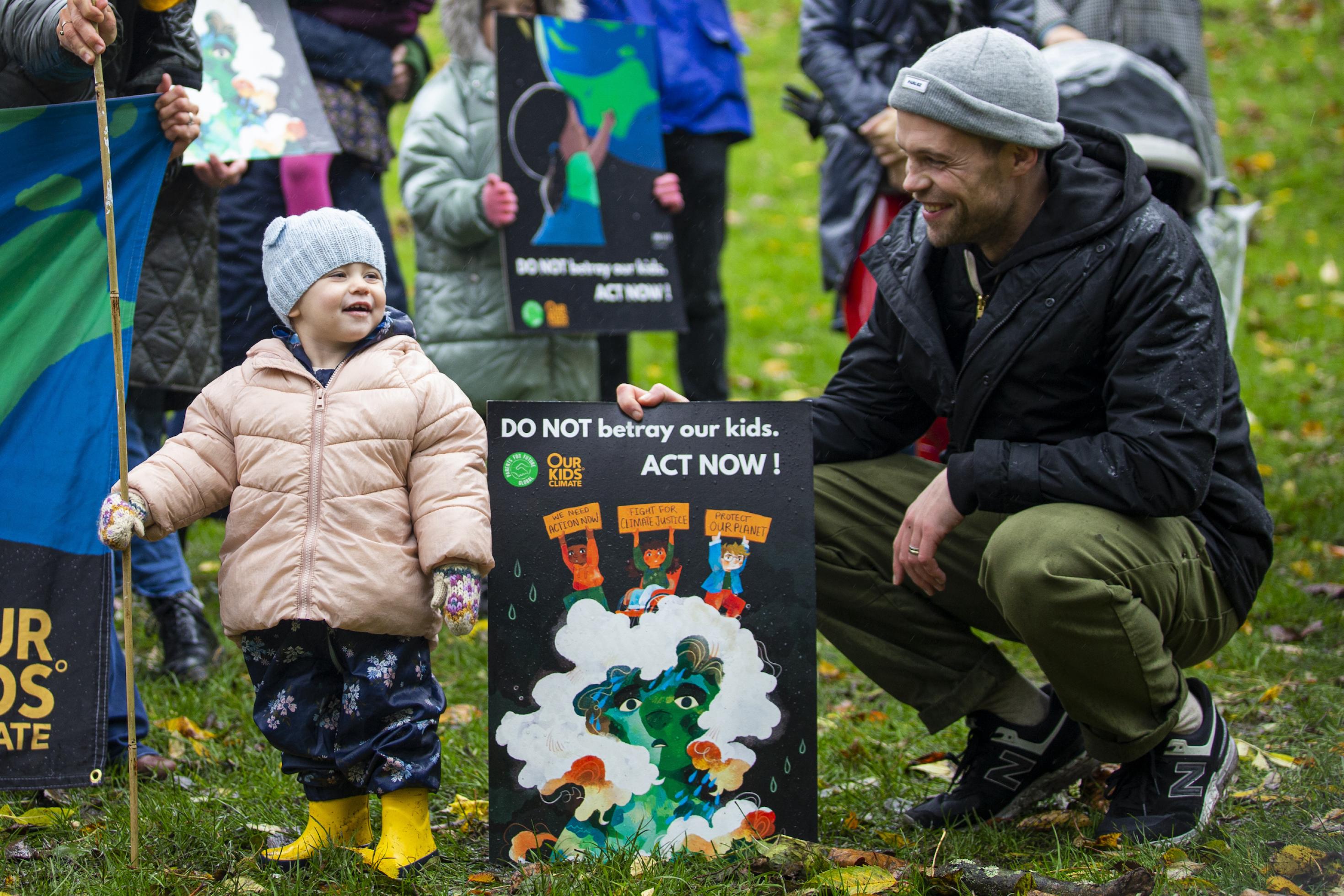 Ein lächelndes Kleinkind neben einem jungen Mann, der ein Protestschild hält. Auf dem Schild steht auf Englisch: Verratet unsere Kinder nicht.