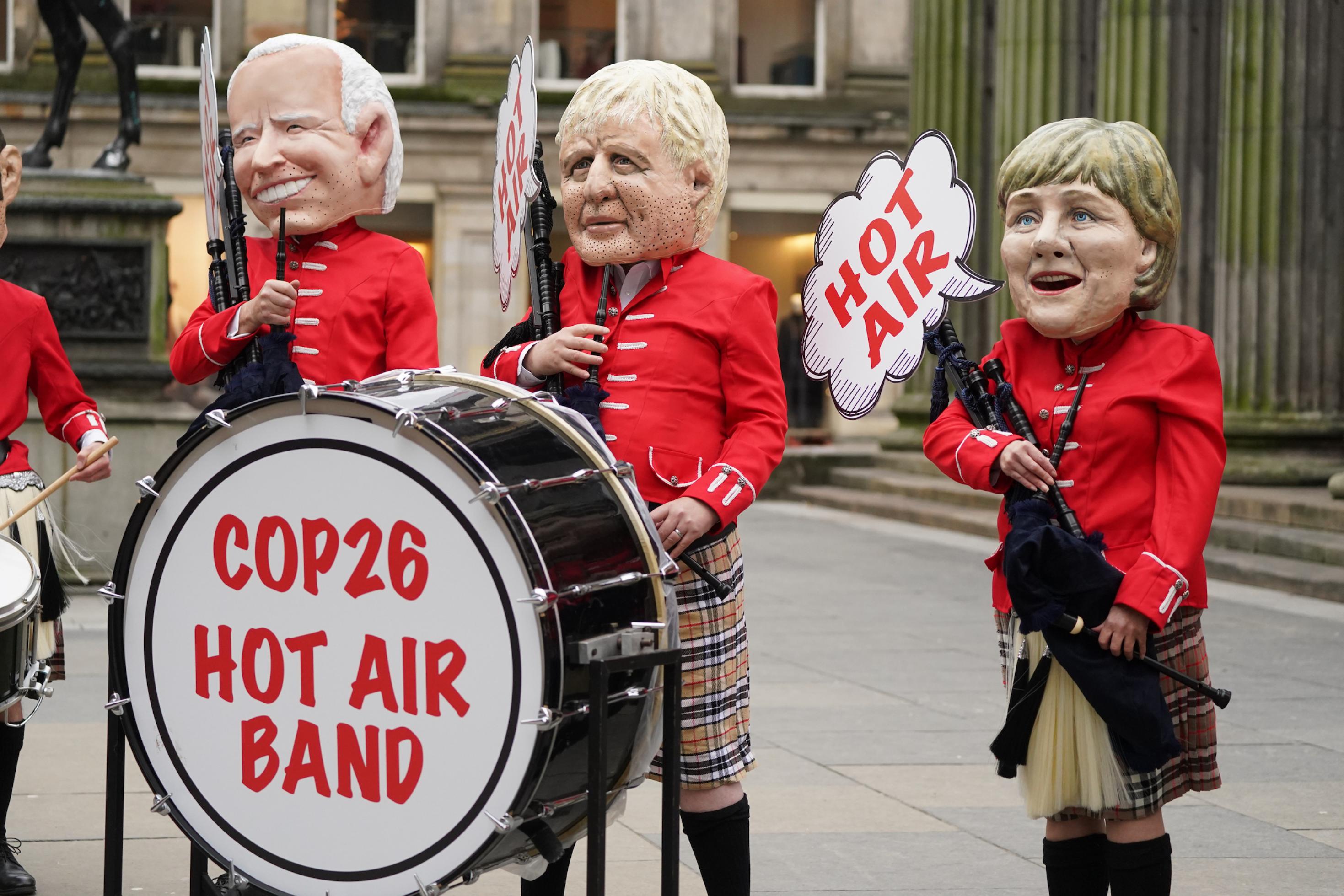 Eine Fake-Band mit Schottenröcken und Pappmacheegesichtern von Biden, Johnson und Merkel und ein Schild mit dem Slogan „Hot Air Band“.