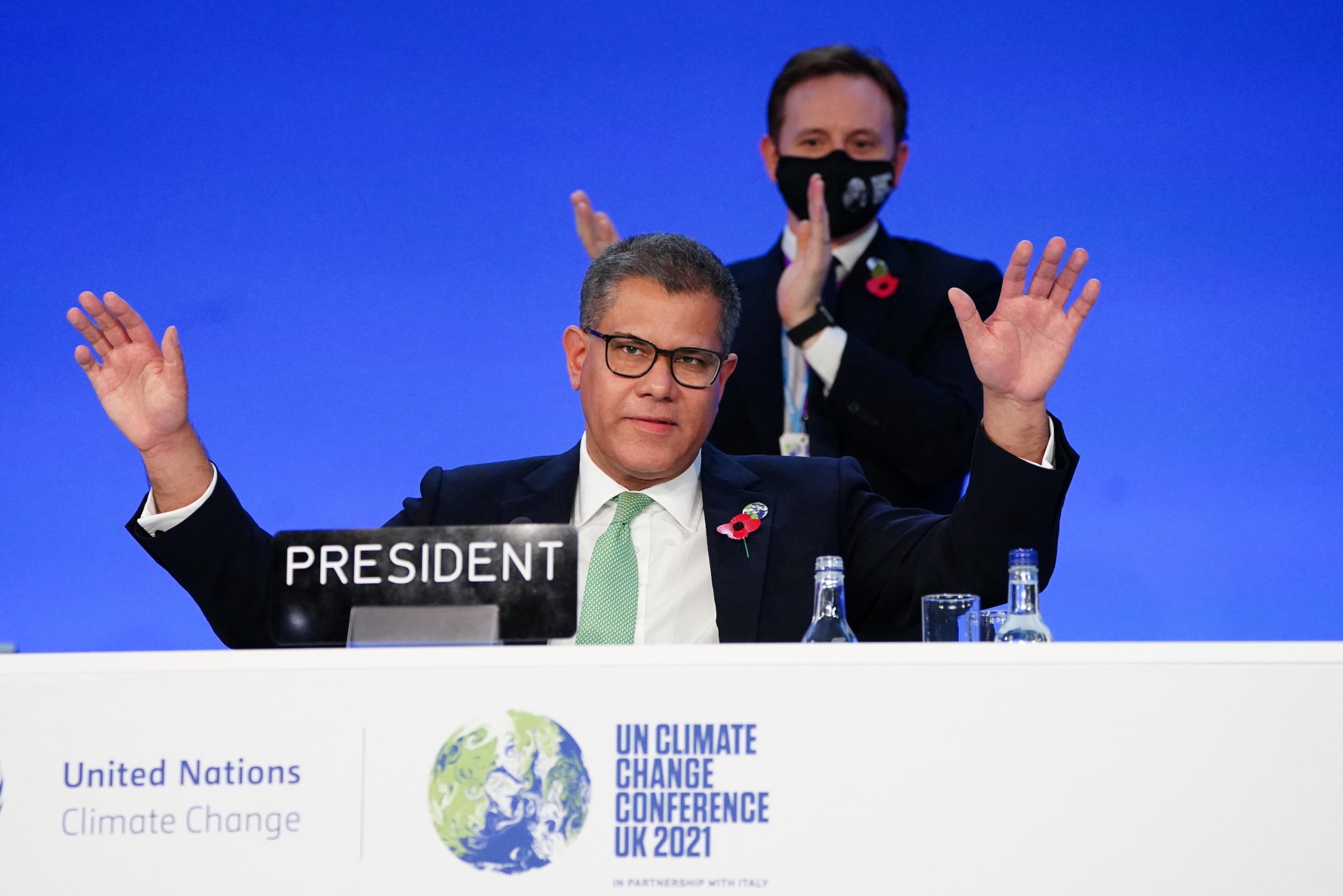 Sharma, ein mittelalter Mann mit Brille, breitet seine Hände vor dem Publikum aus, im Hintergrund applaudiert ihm ein Mann.