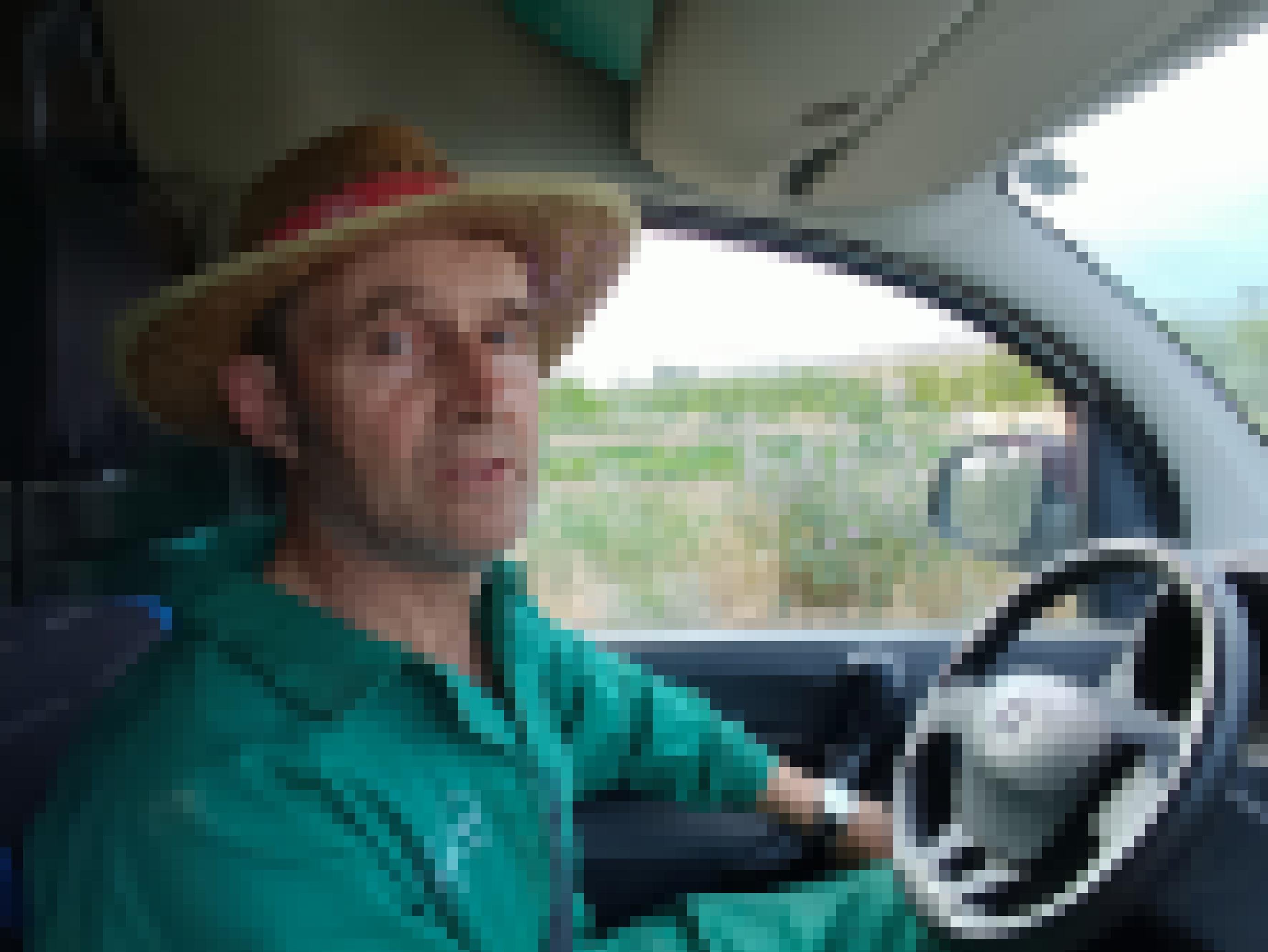 Ein Mann im grünen Overall und mit Strohhut sitzt in einem Auto und blickt fragend in die Kamera.