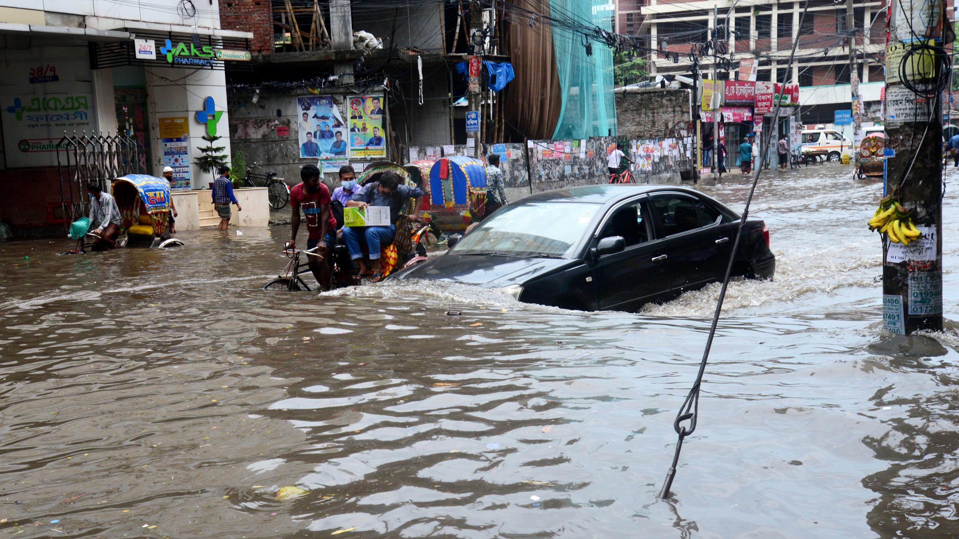 Eine überflutete Straße in Dhaka, Bangladesch, 2020. Autos, Radfahrer, Fußgänger kämpfen sich durch die Wassermassen.