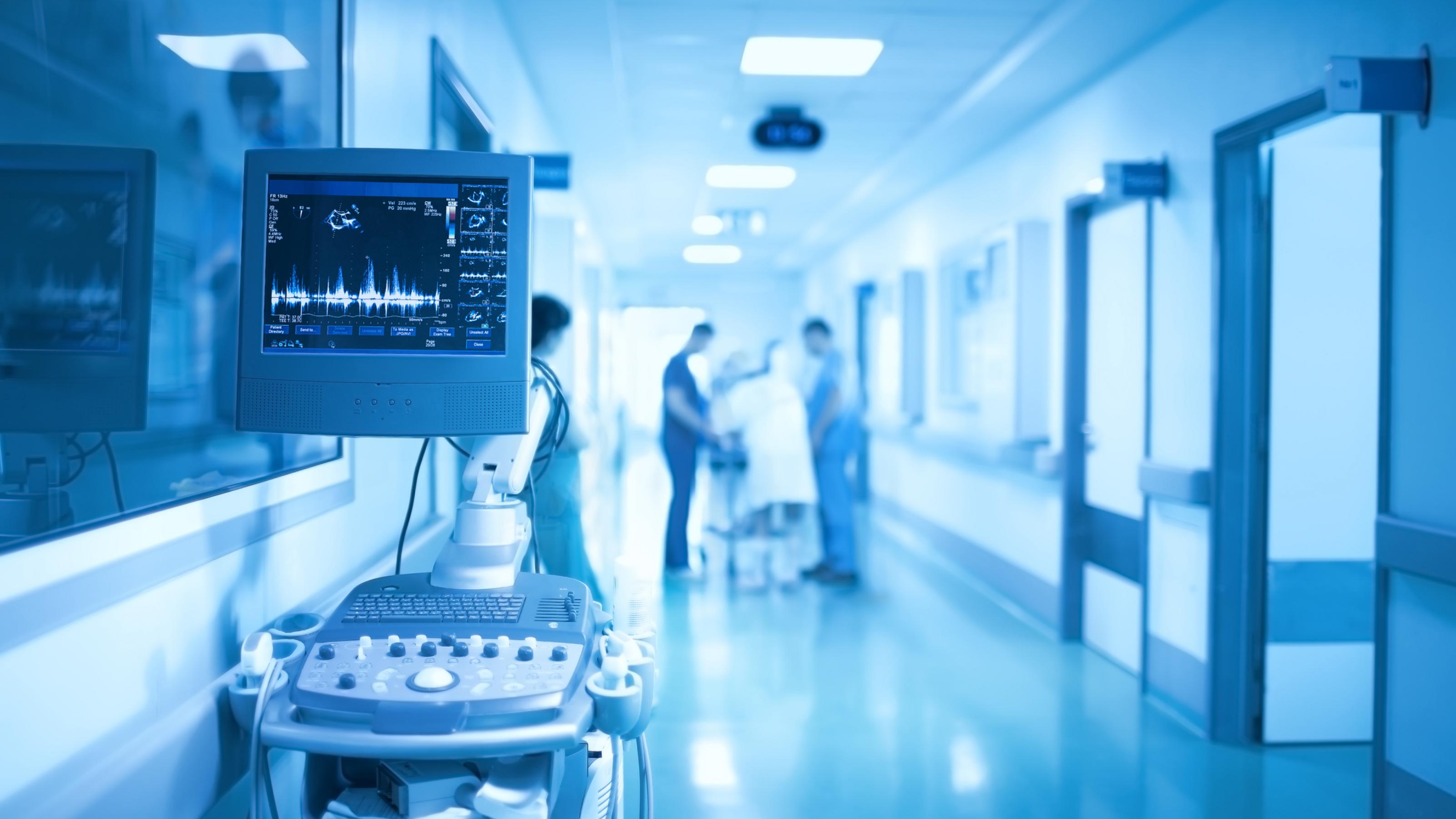 In einem in kalten, blauen Tönen gehaltenen Krankenhausflur stehen im Vordergrund medizinische Gräte, im Hintergrund verschwommen Personal
