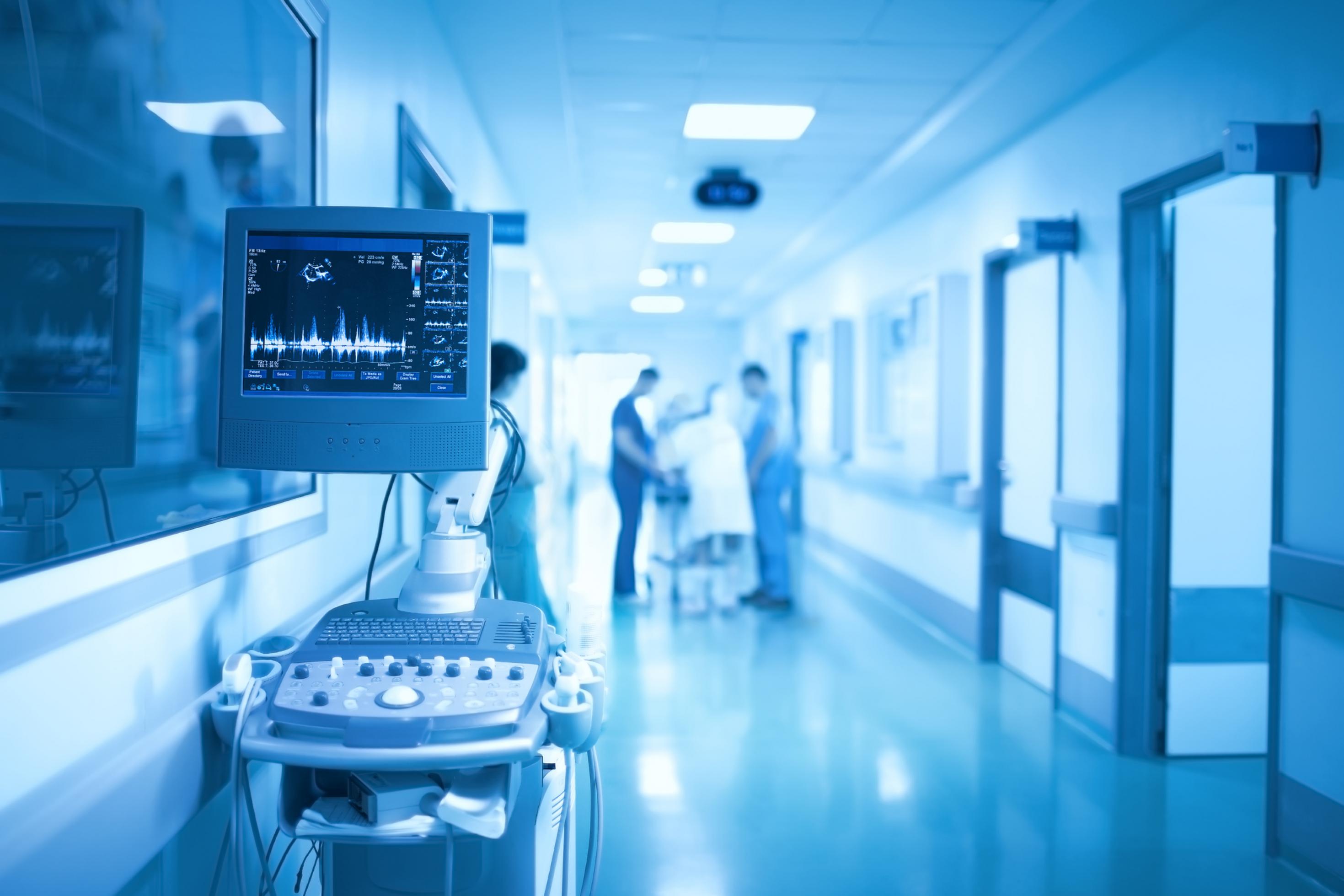 In einem in kalten, blauen Tönen gehaltenen Krankenhausflur stehen im Vordergrund medizinische Gräte, im Hintergrund verschwommen Personal
