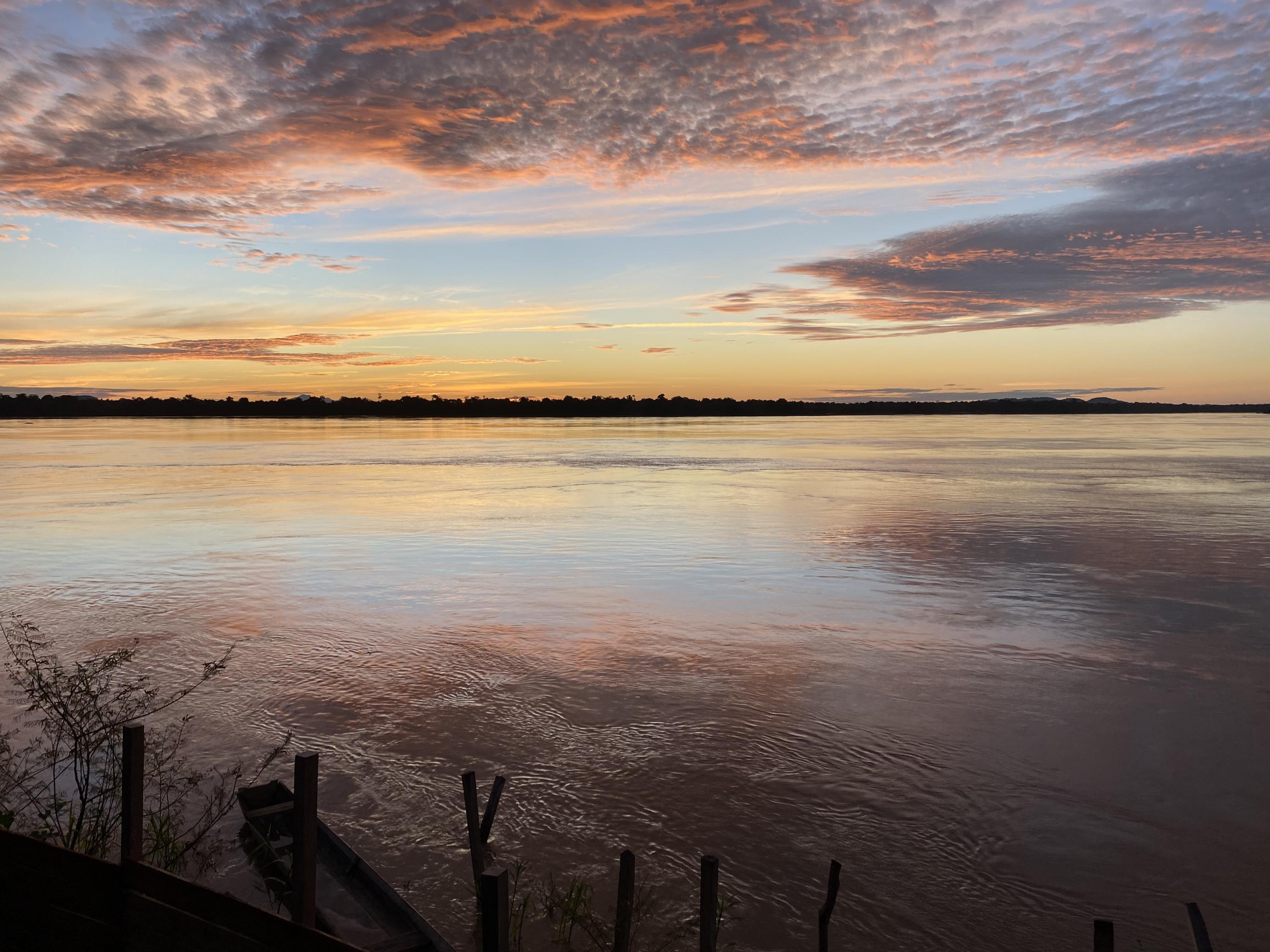 Dunkelrot-orangen-rosafarbene Wolken, die sich im Fluss spiegeln, im Vordergrund ein halb gesunkenes Holzboot.